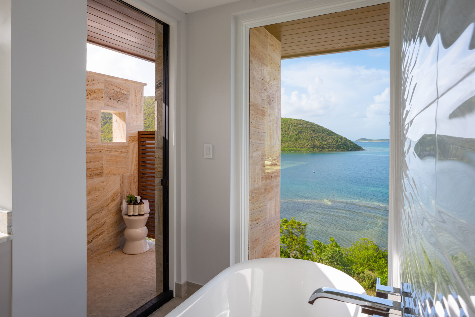 A modern bathroom in Deep Bay Villa features a white bathtub with scenic coastal views. An open door reveals an outdoor shower area enclosed by stone walls. Large windows offer glimpses of the ocean and lush green hills, cultivating a serene, tropical ambiance that epitomizes villa luxury.