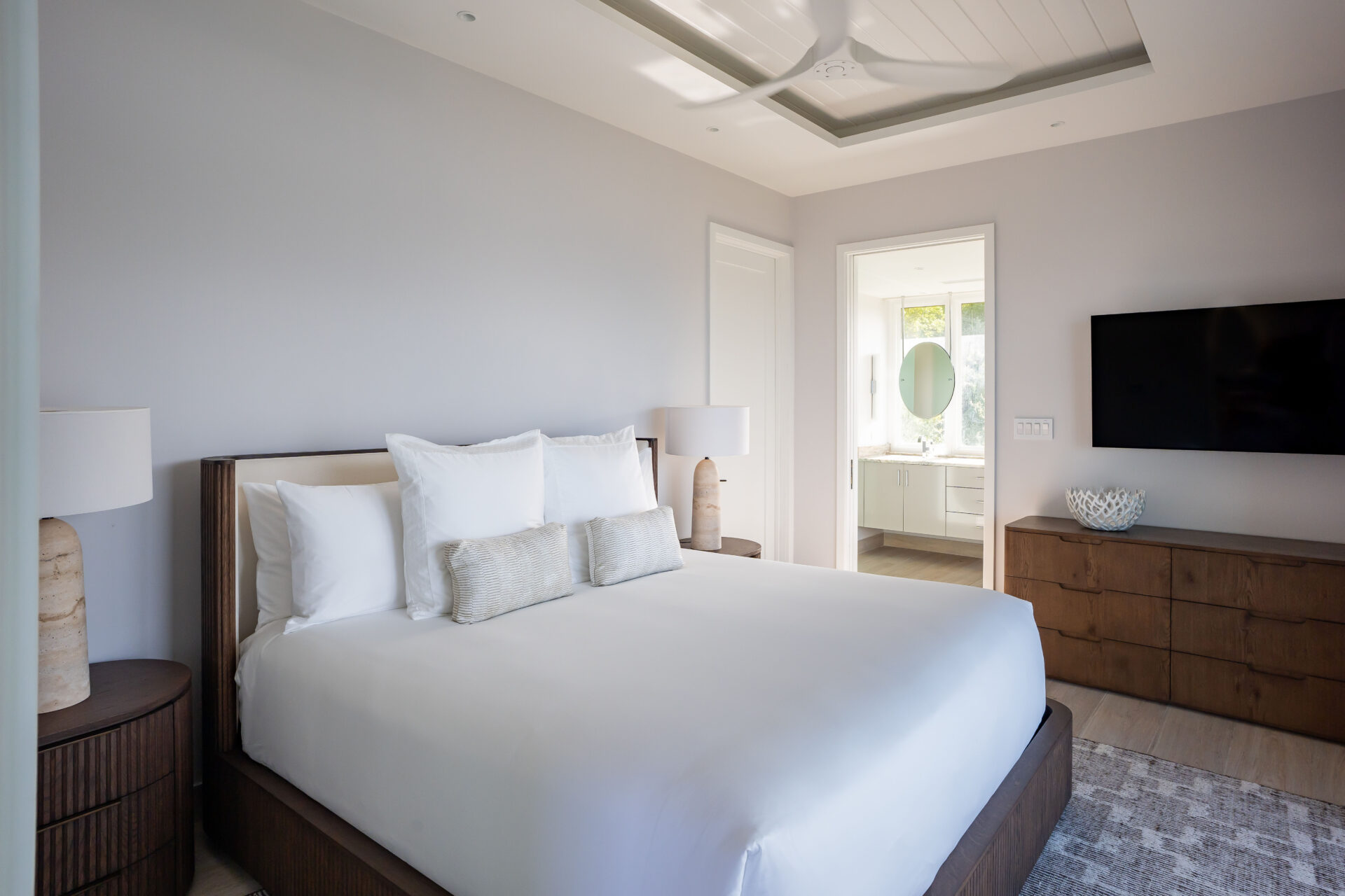 A modern bedroom at Deep Bay Villa features a large bed with white linens and multiple pillows. Two wooden nightstands with lamps flank the bed. A flat-screen TV is mounted on the wall above a wooden dresser, and a doorway leads to a bright bathroom.