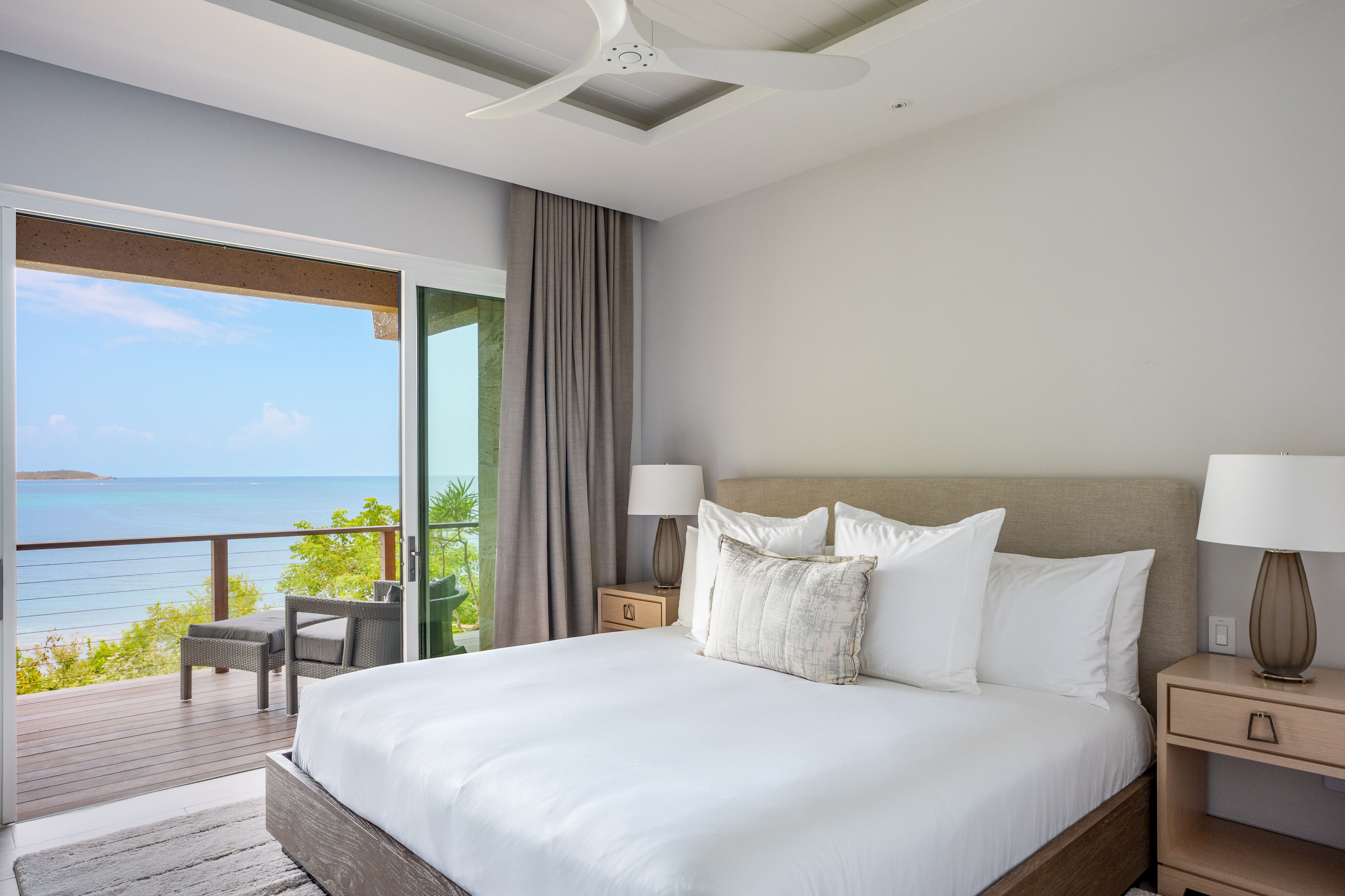 A bright, modern bedroom at Deep Bay Villa features a large bed with white linens. Two bedside tables with lamps flank the bed. Glass doors open to a balcony overlooking Deep Bay and the vast blue sky. Neutral colors and a ceiling fan enhance the serene atmosphere.