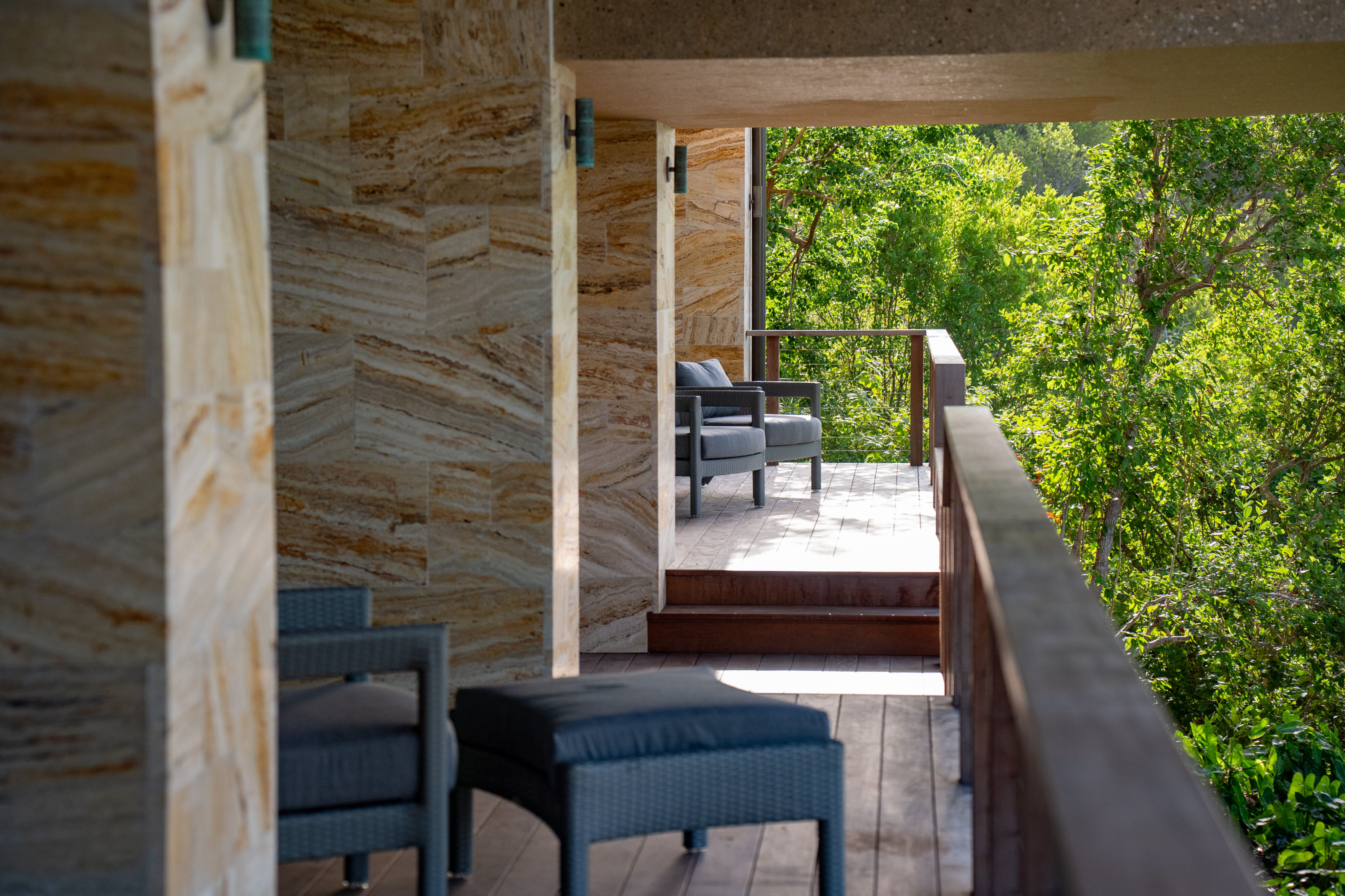 The sunlit balcony of Deep Bay Villa, featuring wicker furniture like a chair and ottoman, overlooks lush green trees. The stone-textured walls and wooden floor add to the serene ambiance of Villa DB1's natural setting.