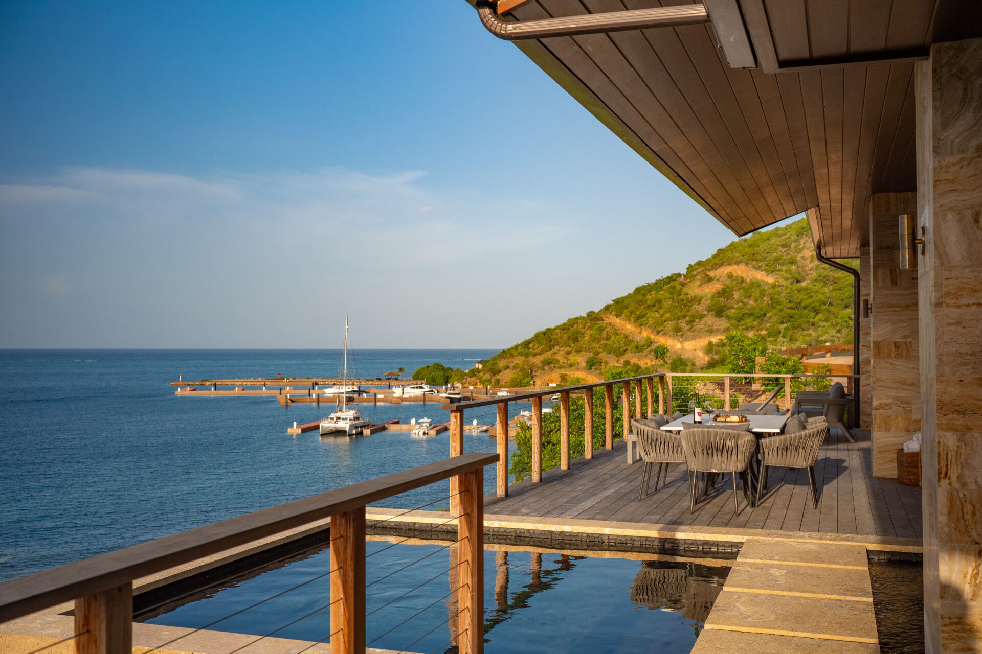 Discover the elegance of Deep Bay Villa's modern terrace, overlooking a serene bay with sailboats and a green hillside under a clear blue sky. Enjoy dining by the pool, framed by stylish wooden railings, and soak in the tranquil vibes of this exquisite DB1 retreat.