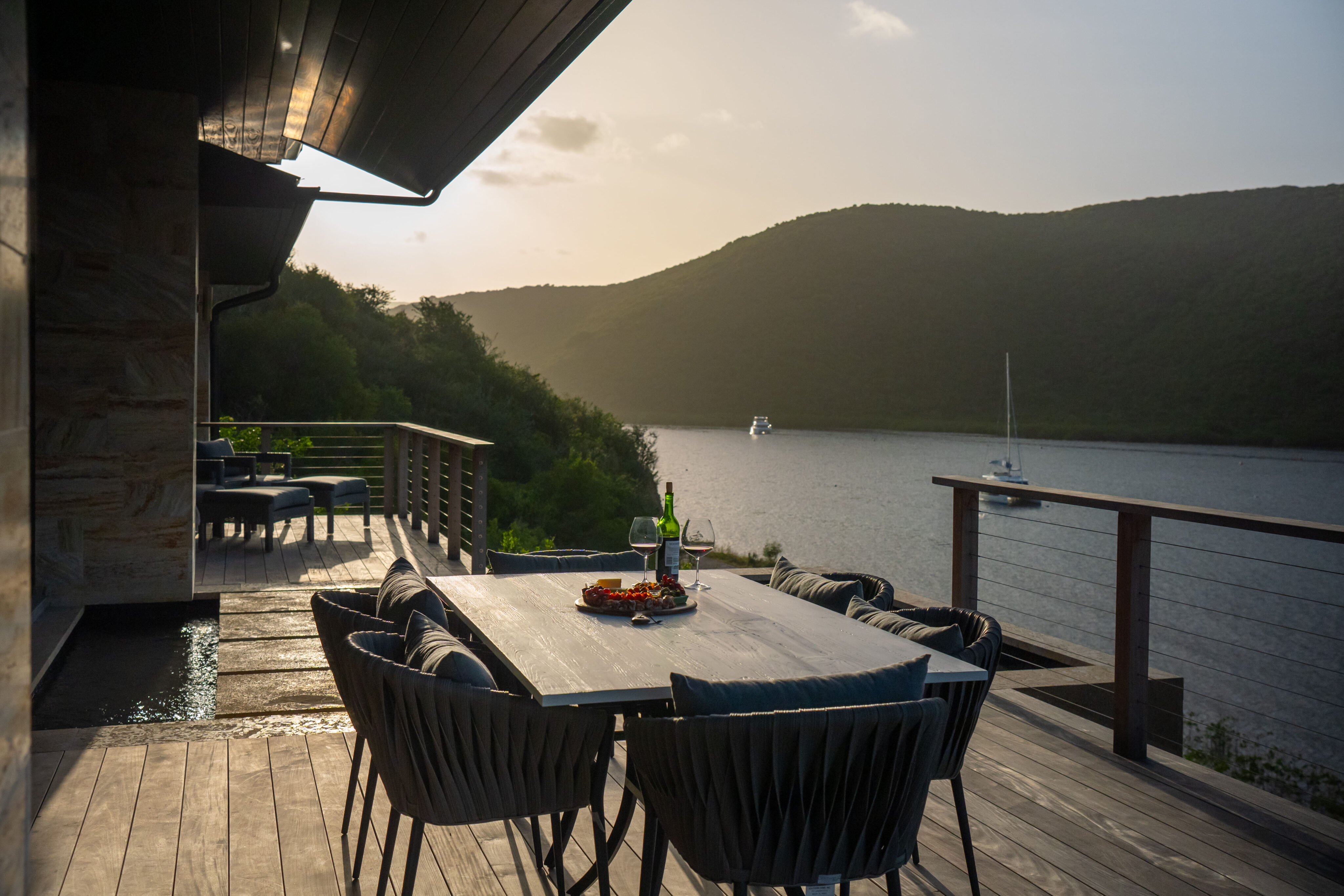 A dining table set for two on the wooden deck of Deep Bay Villa overlooks a tranquil lake at sunset. A bottle of wine and glasses are on the table, surrounded by chairs. Hills rise in the background, with a few boats gently drifting across the water.