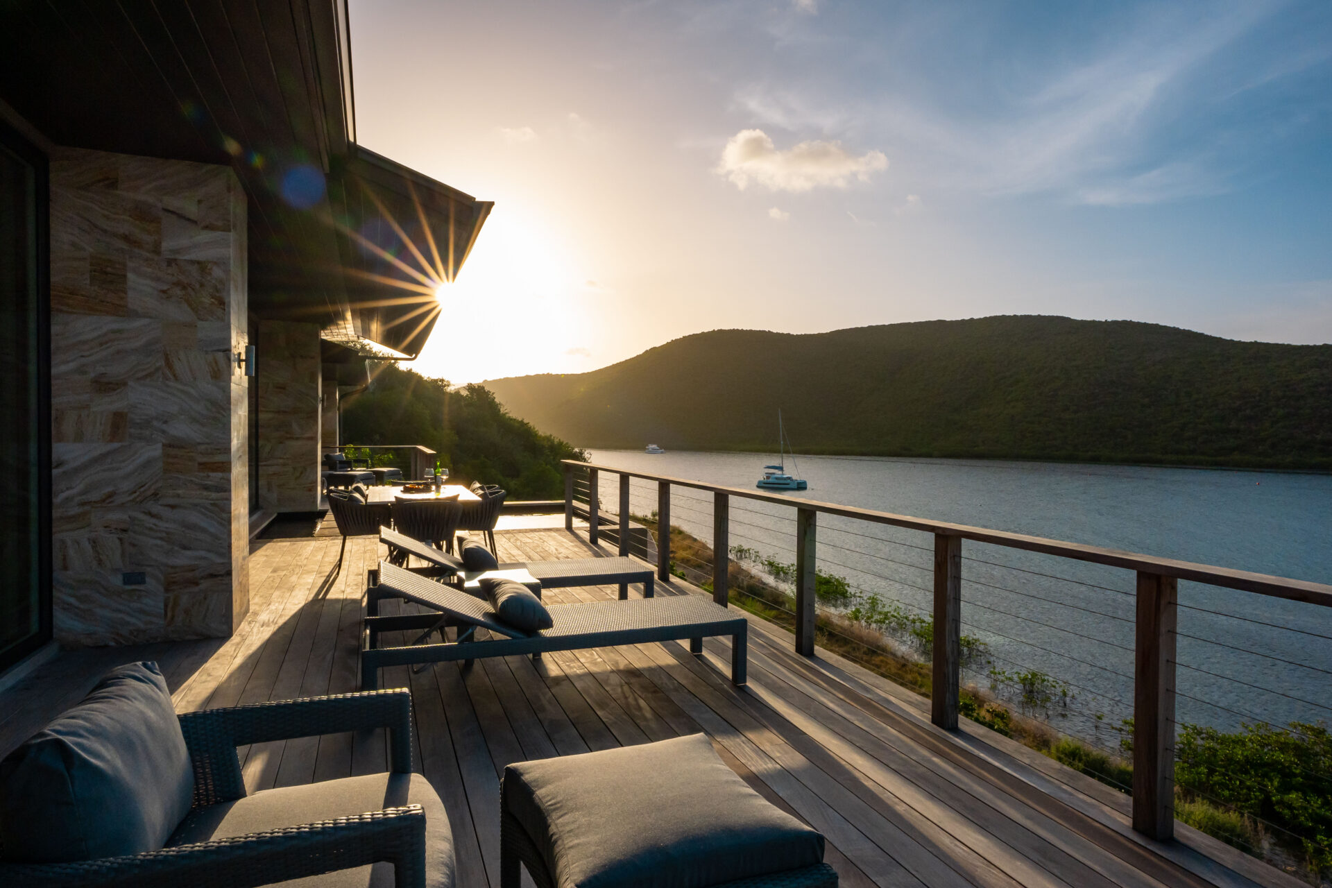 Perched on the wooden deck of Deep Bay Villa, the sunset view over the tranquil lake is mesmerizing. Lounge chairs await as you take in the surrounding hills, while a boat gently sways. Sunlight dances with a lens flare, painting an unforgettable evening at DB1.