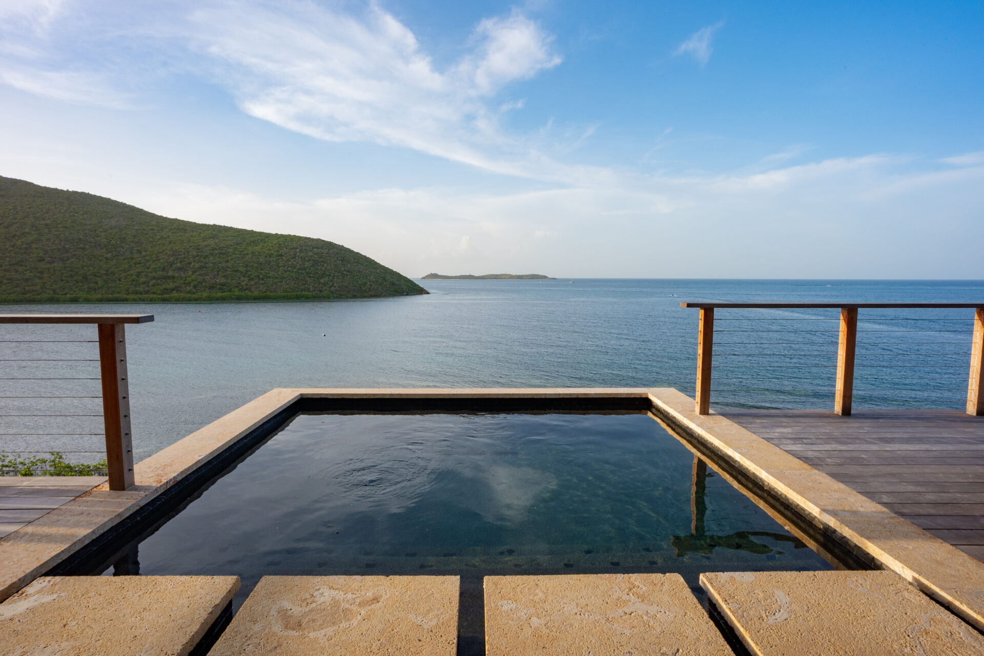 A small, serene pool at the Villa overlooks Deep Bay's tranquil ocean, with lush green hills in the background. The scene is set under a bright blue sky scattered with a few white clouds. A wooden railing borders the edge of the deck.