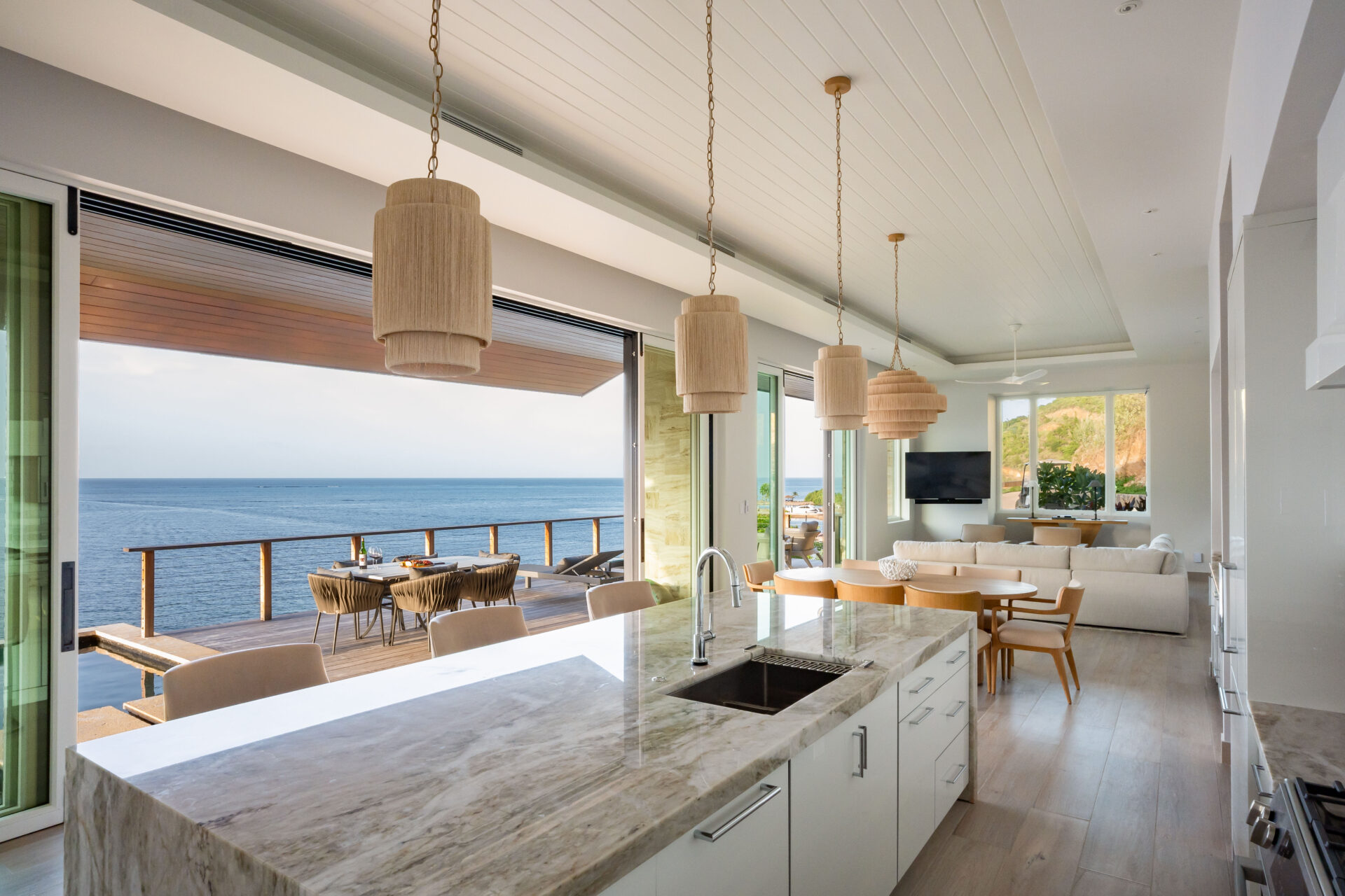 The Deep Bay Villa boasts a modern open-concept kitchen and living area with large windows offering captivating ocean views. This exquisite space features a marble island, pendant lights, and a dining table. Outside, the balcony with a dining set overlooks the serene waters of Deep Bay.