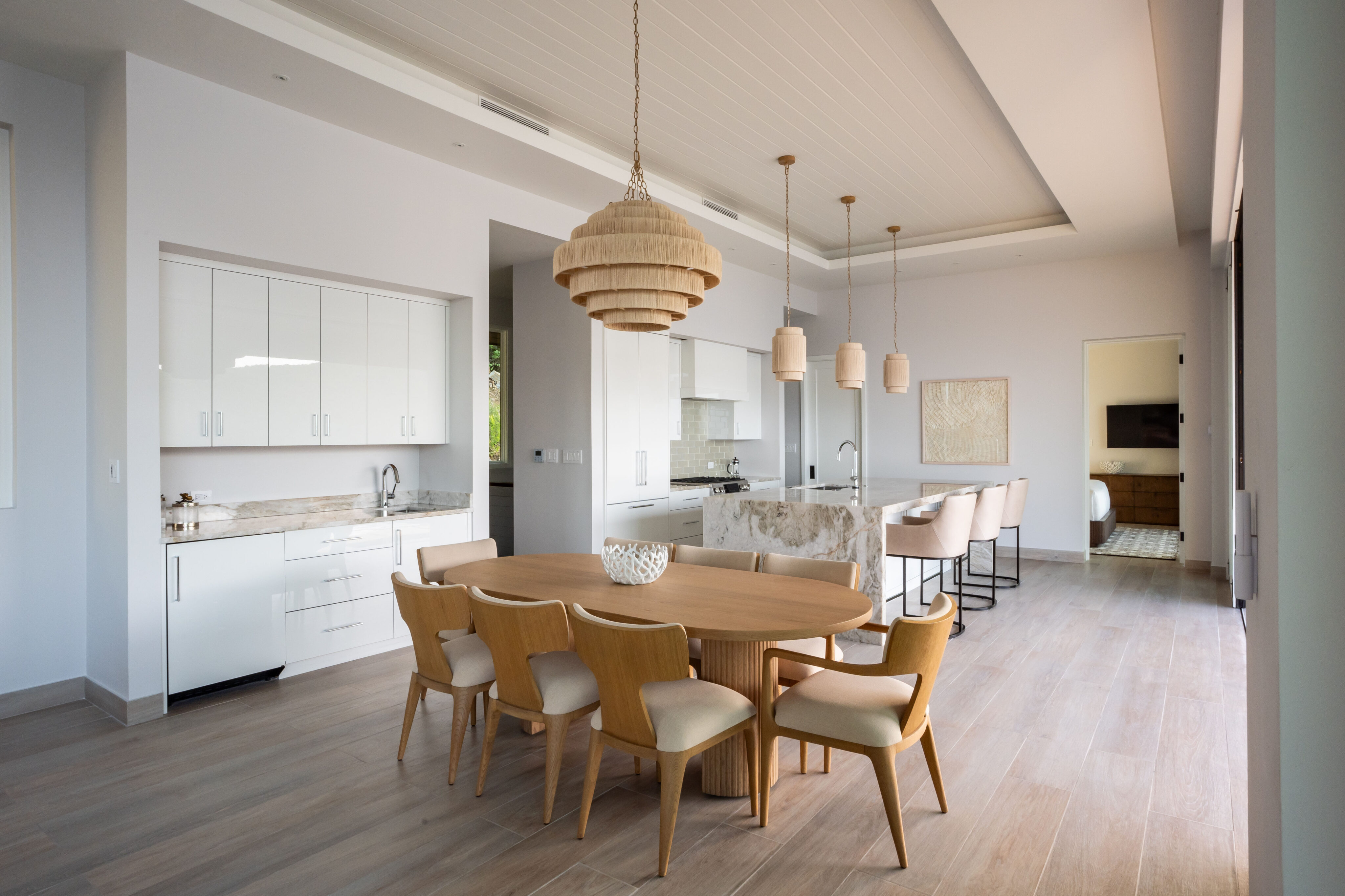 A modern kitchen and dining area with light wood flooring, a large wooden dining table surrounded by six chairs, and a set of pendant lights above. The Villa features a kitchen island with bar stools, white cabinets, and a neutral color palette reminiscent of Deep Bay's serene elegance.