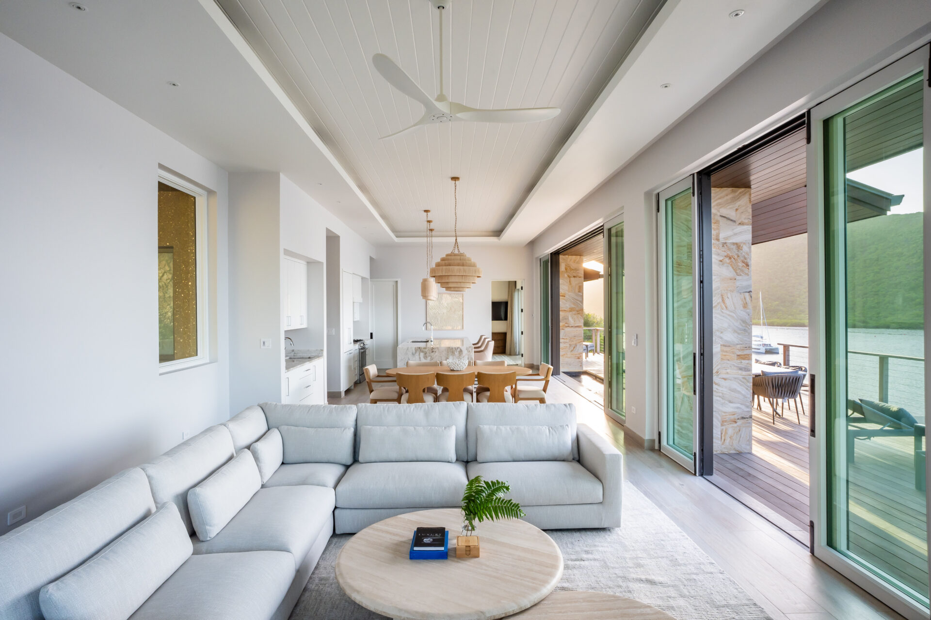 A bright, modern living room at Villa DB1 features a large sectional sofa and a round coffee table topped with books and a plant. Sliding glass doors open to a patio with a lake view. A dining area and kitchen are visible in the background, inviting guests to enjoy the comfort of Deep Bay Villa.