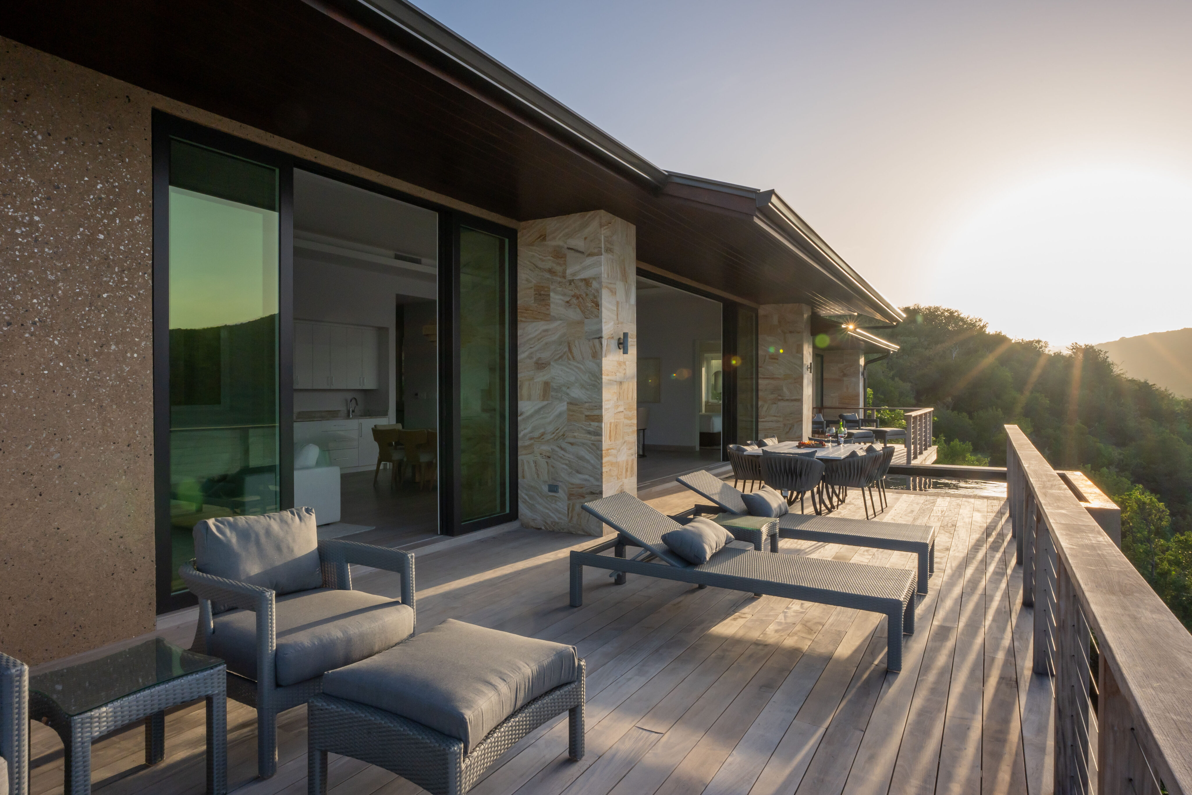 A modern terrace at sunset features cushioned lounge chairs and a dining table. Overlooking the lush green landscape, Deep Bay Villa showcases the sun setting in the background, casting warm light over its sleek, minimalist design.