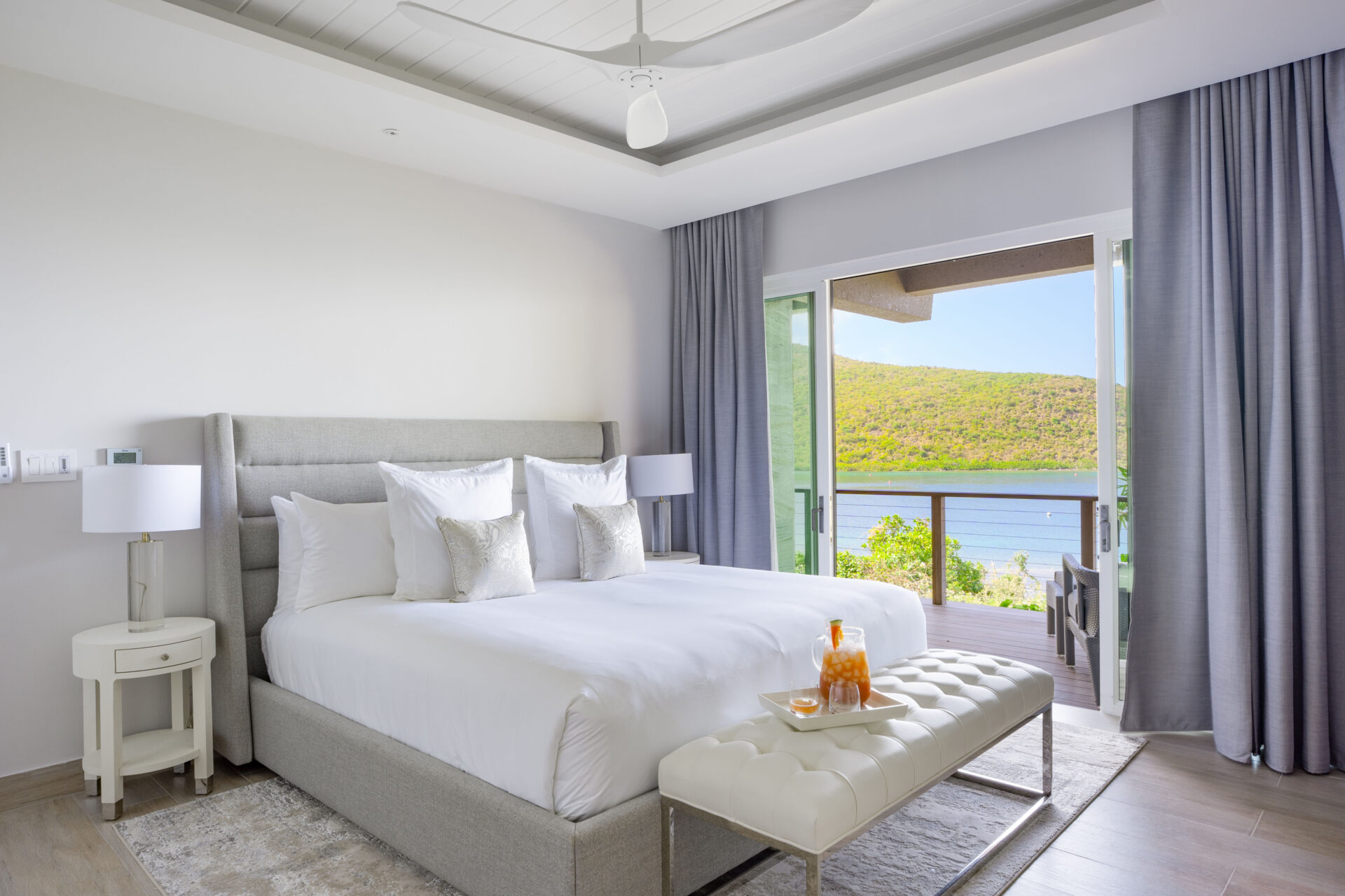 In the modern bedroom of Deep Bay Villa, a large bed with white linens and decorative pillows dominates the space. A cushioned bench rests at its foot, flanked by two lamps. Glass doors open to a balcony offering stunning views of hills and the serene waters of Deep Bay.