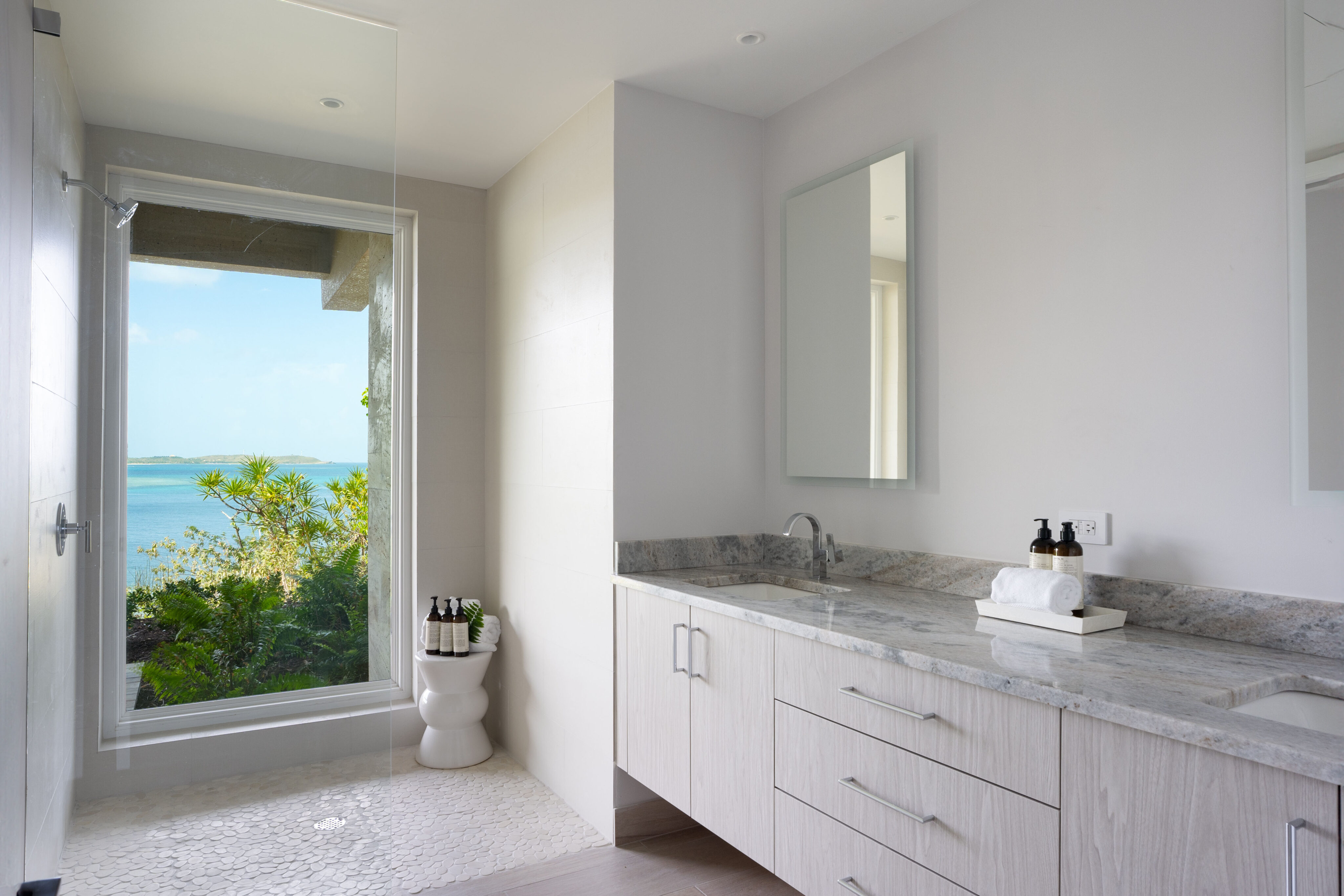 A modern bathroom in Deep Bay Villa showcases light wood cabinetry and a large mirror. The bright, airy space features a shower with a glass door, offering views of the ocean and lush greenery outside the window—a true DB1 sanctuary.