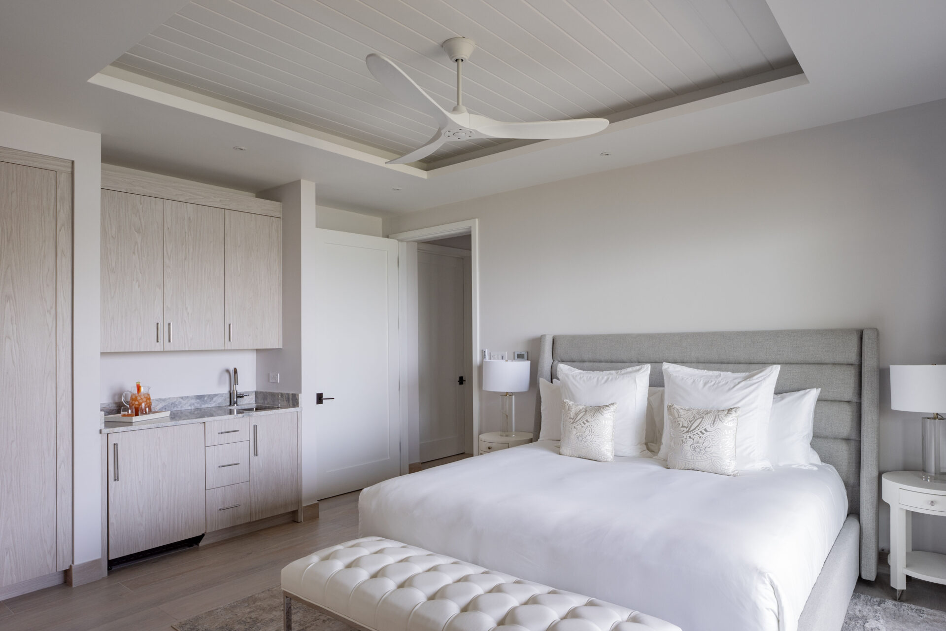 A modern bedroom in Deep Bay Villa features a large bed with white linens, flanked by two white nightstands with lamps. The room includes a ceiling fan and a small kitchenette with light wood cabinets and a gray countertop. A padded bench sits at the foot of the bed, perfect for relaxing in the serene setting.