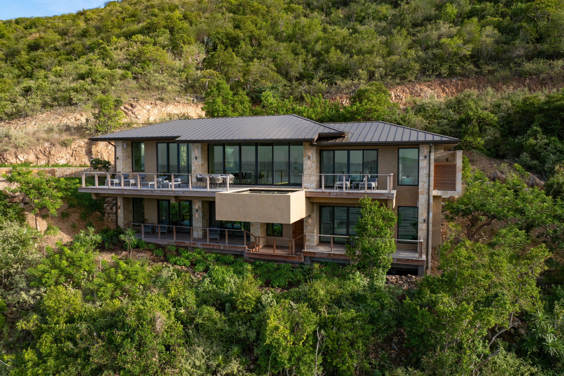 Nestled on a lush, green hillside surrounded by trees, Deep Bay Villa is a modern two-story house with large glass windows and balconies. Its flat roof seamlessly integrates with the natural setting.