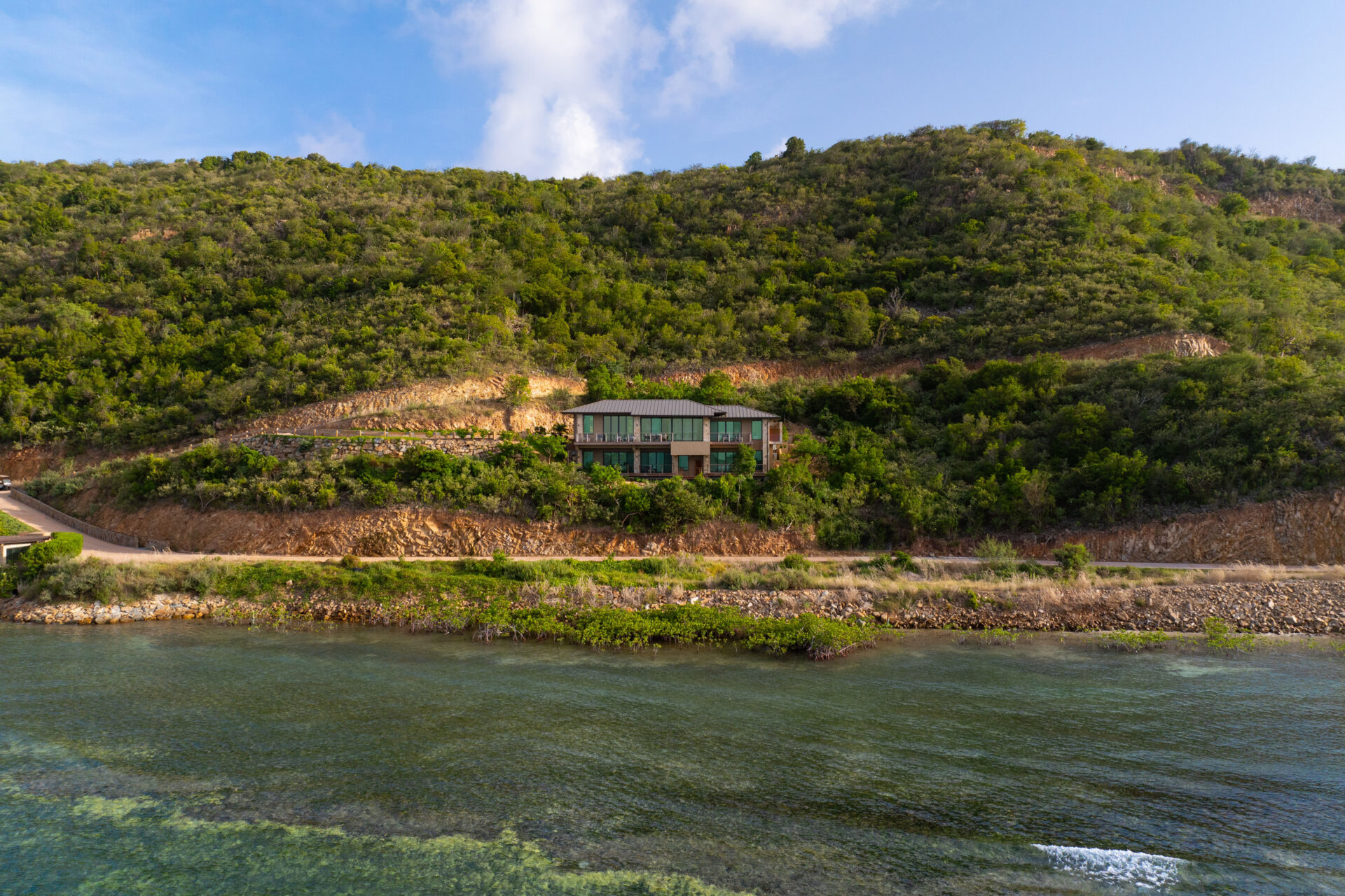 A secluded villa with a green roof nestled into a lush hillside, overlooking the clear, calm waters of Deep Bay. The surrounding area is covered in dense greenery, with a road running along the edge of the water.