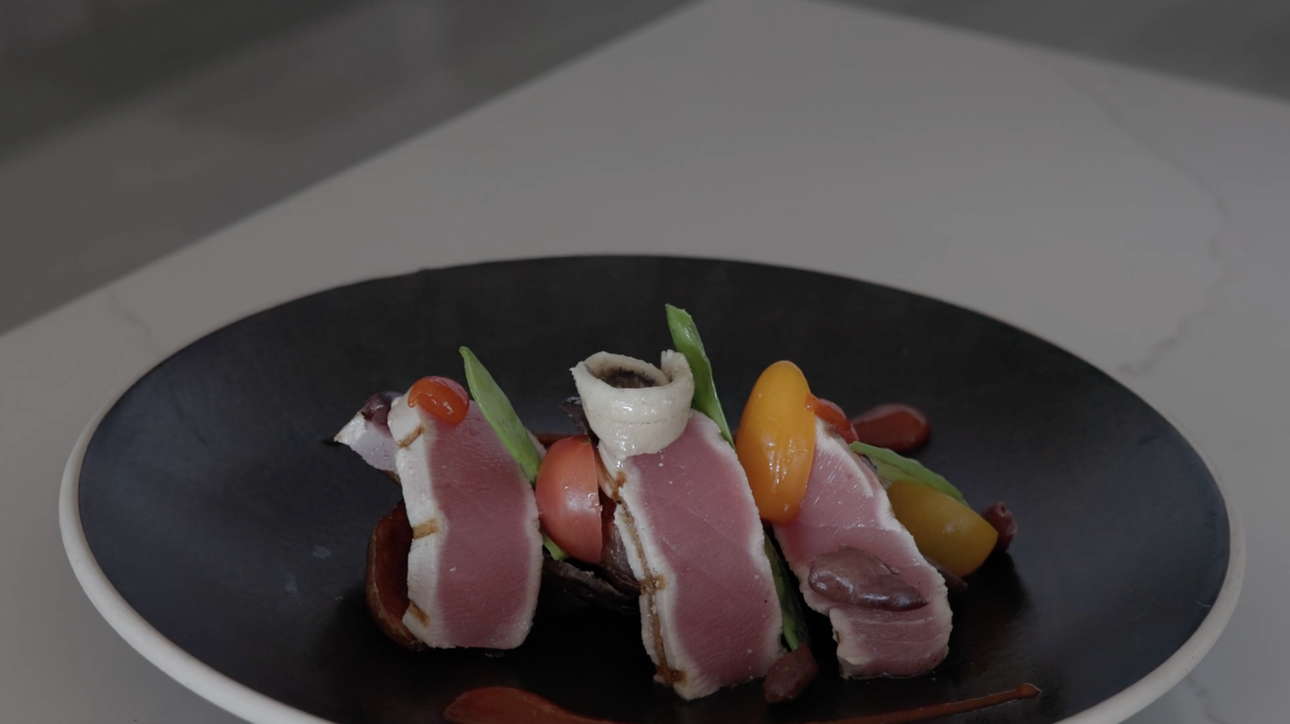 A gourmet dining dish featuring slices of seared tuna, garnished with green beans, cherry tomatoes, and a rolled anchovy, beautifully arranged on a black plate with artful drizzles of sauce. The plate sits elegantly on a white surface.