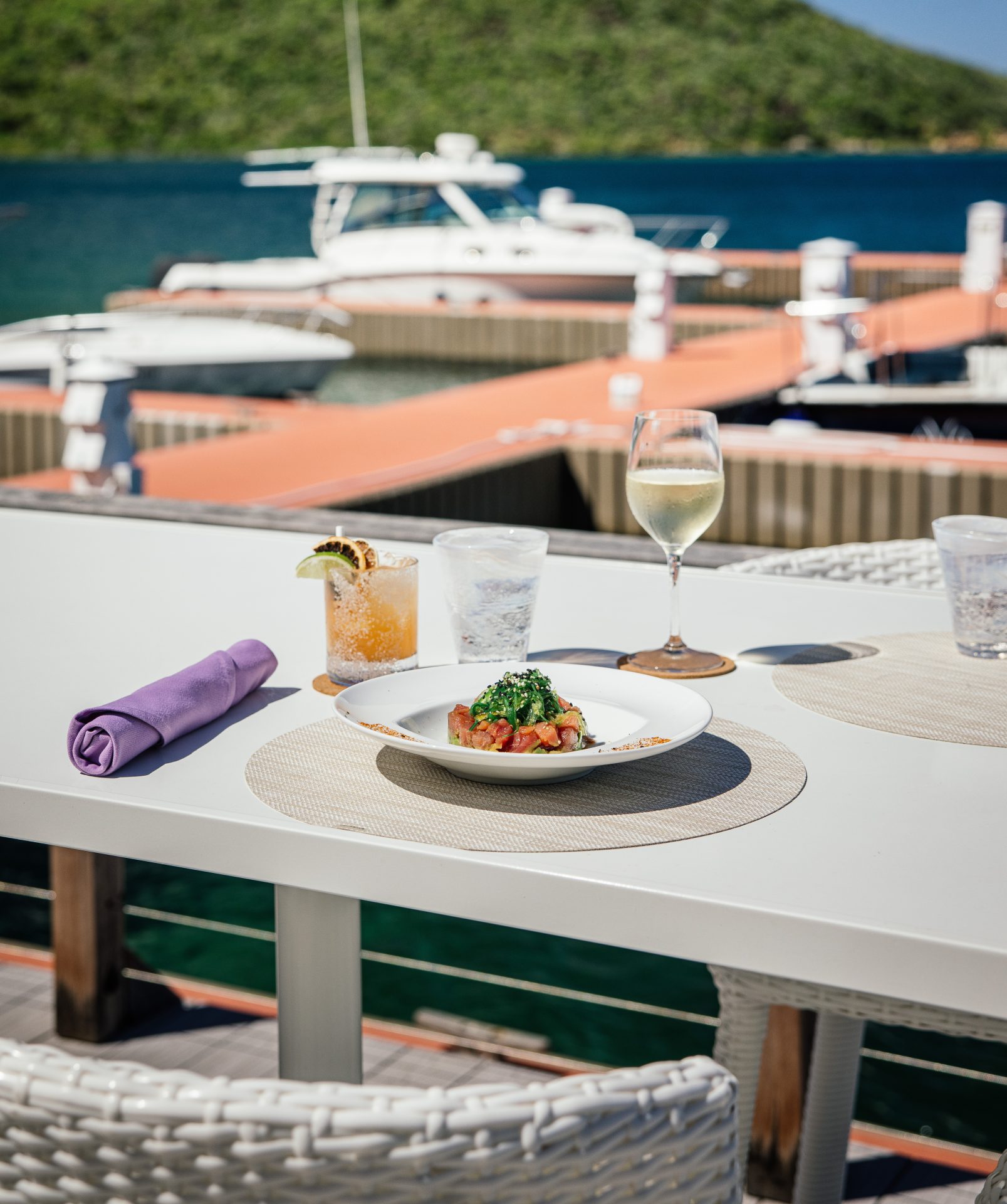 A white outdoor dining table set with a plate of food, two glasses—one with a cocktail and one with white wine—and a purple napkin. The table overlooks the Nova Marina, where a yacht is docked against picturesque lush green hills in the background.