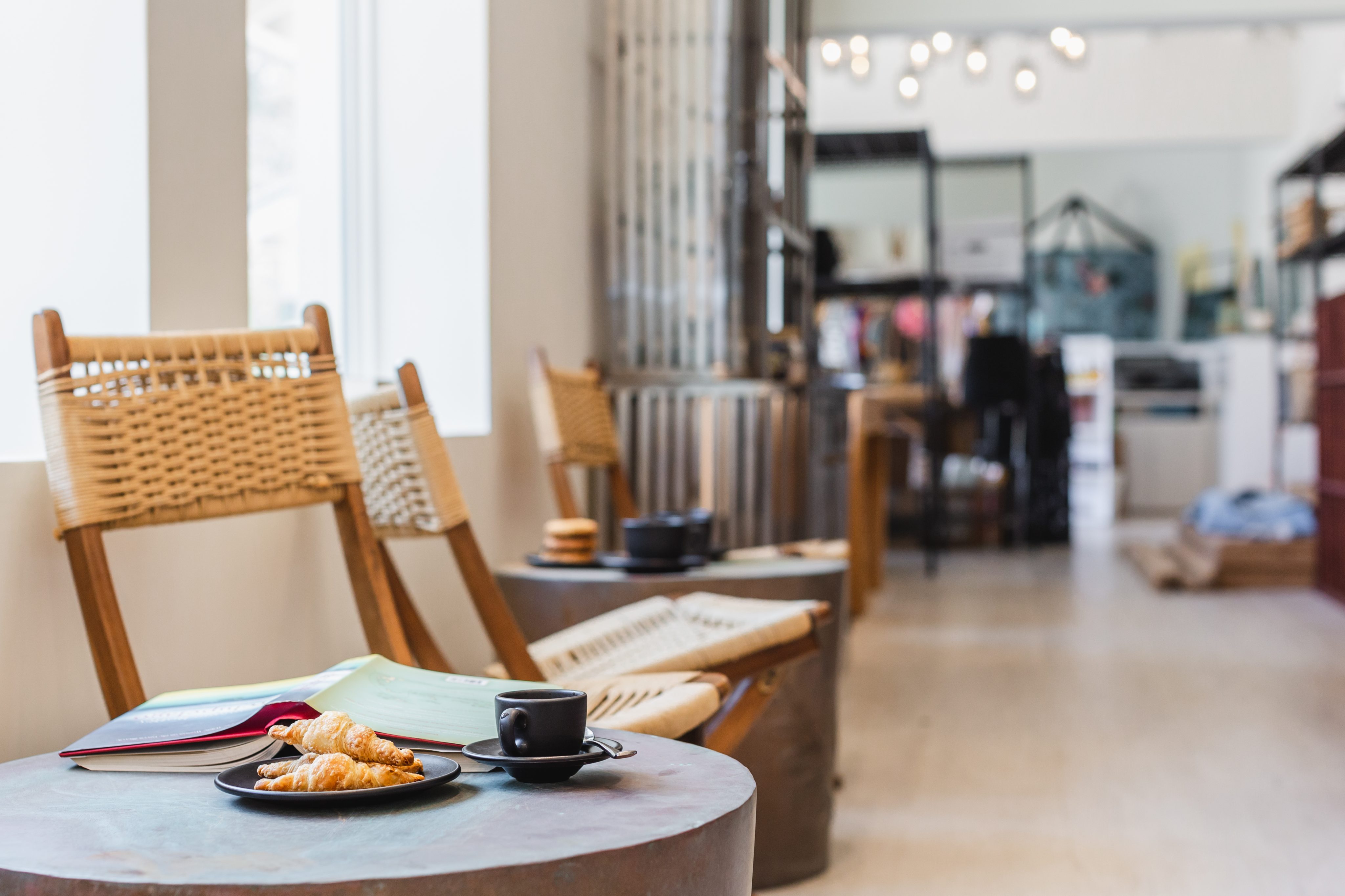 A cozy coffee shop interior with wicker chairs along a wall, evoking a marina's relaxed charm. A small table in the foreground holds an open book, a plate with croissants, and a cup of coffee. The background features shelves with various items and hanging lights.