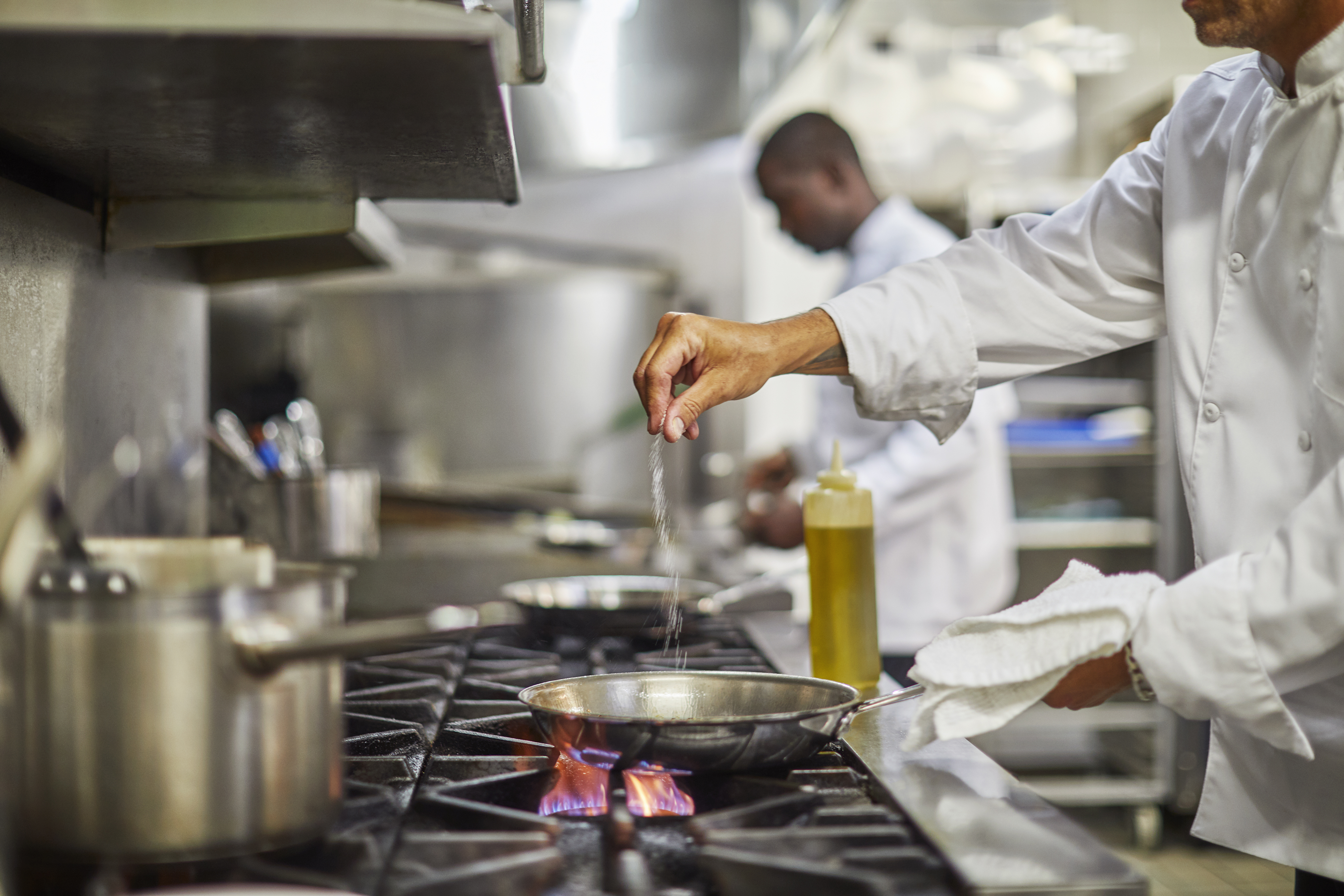 Two chefs work in a professional kitchen. One chef in the foreground seasons a dish in a skillet on the stovetop, embodying the flavors of Island Dining in the British Virgin Islands, while the other chef in the background is focused on preparation work. Cooking pots and utensils are visible on the stovetop.