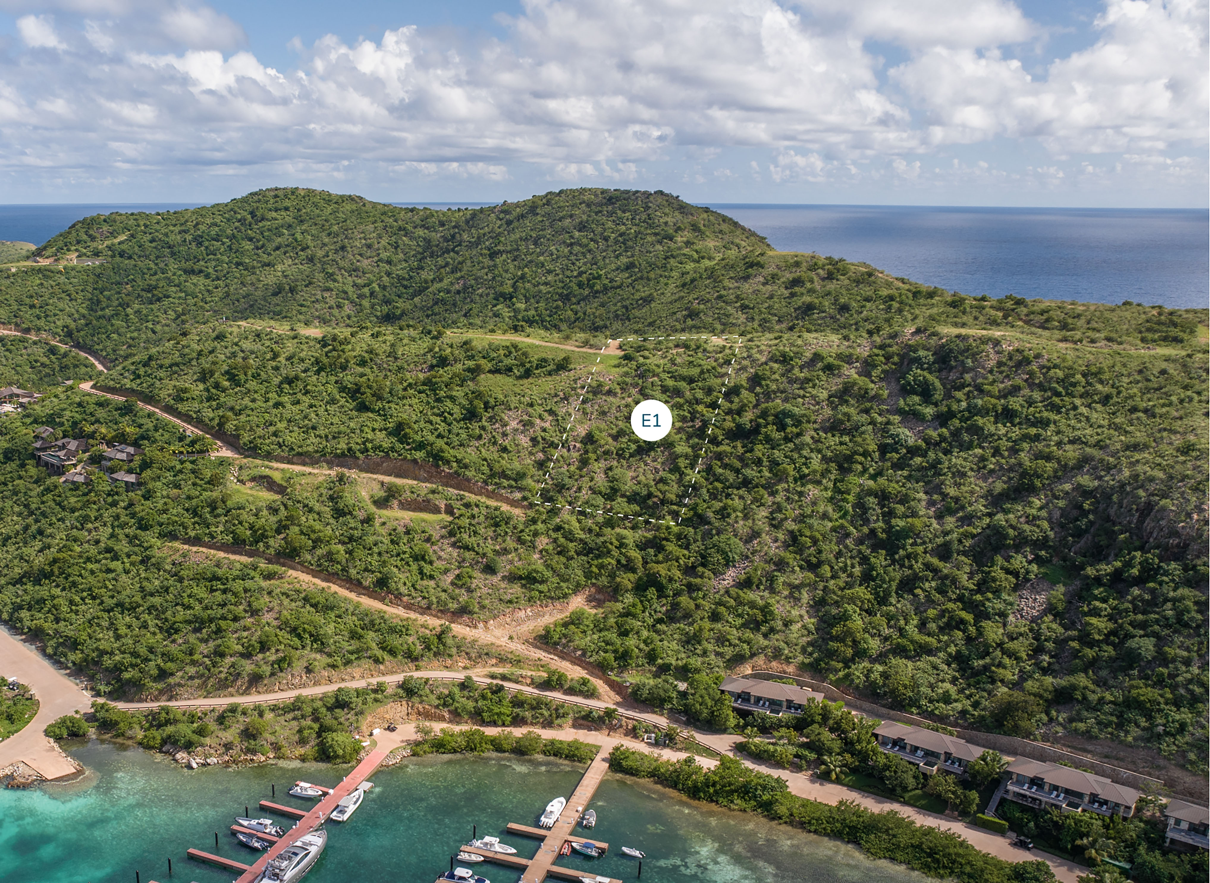 Aerial view of a scenic coastal landscape featuring lush green hills, a marina with boats docked, and several buildings along the waterfront. The area is surrounded by the blue ocean and scattered clouds dot the sky. An "E1" marker indicates an Estate Homesite on one of the hills.