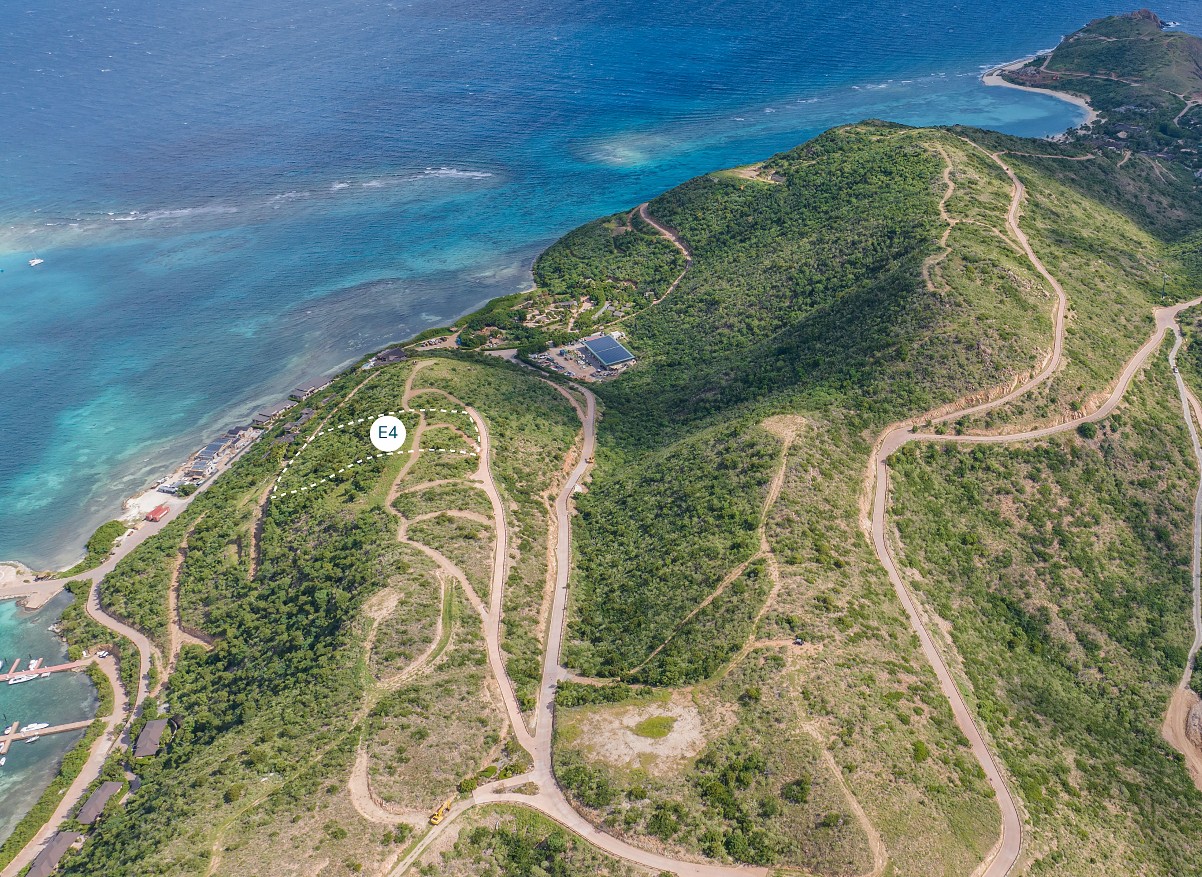 Aerial view of a coastal mountainous region with winding roads zigzagging through green hills. The blue ocean is visible in the background. A white circular marker labeled "E4" is positioned on one of the hills, highlighting a potential homesite. Buildings and a marina are near the shoreline.