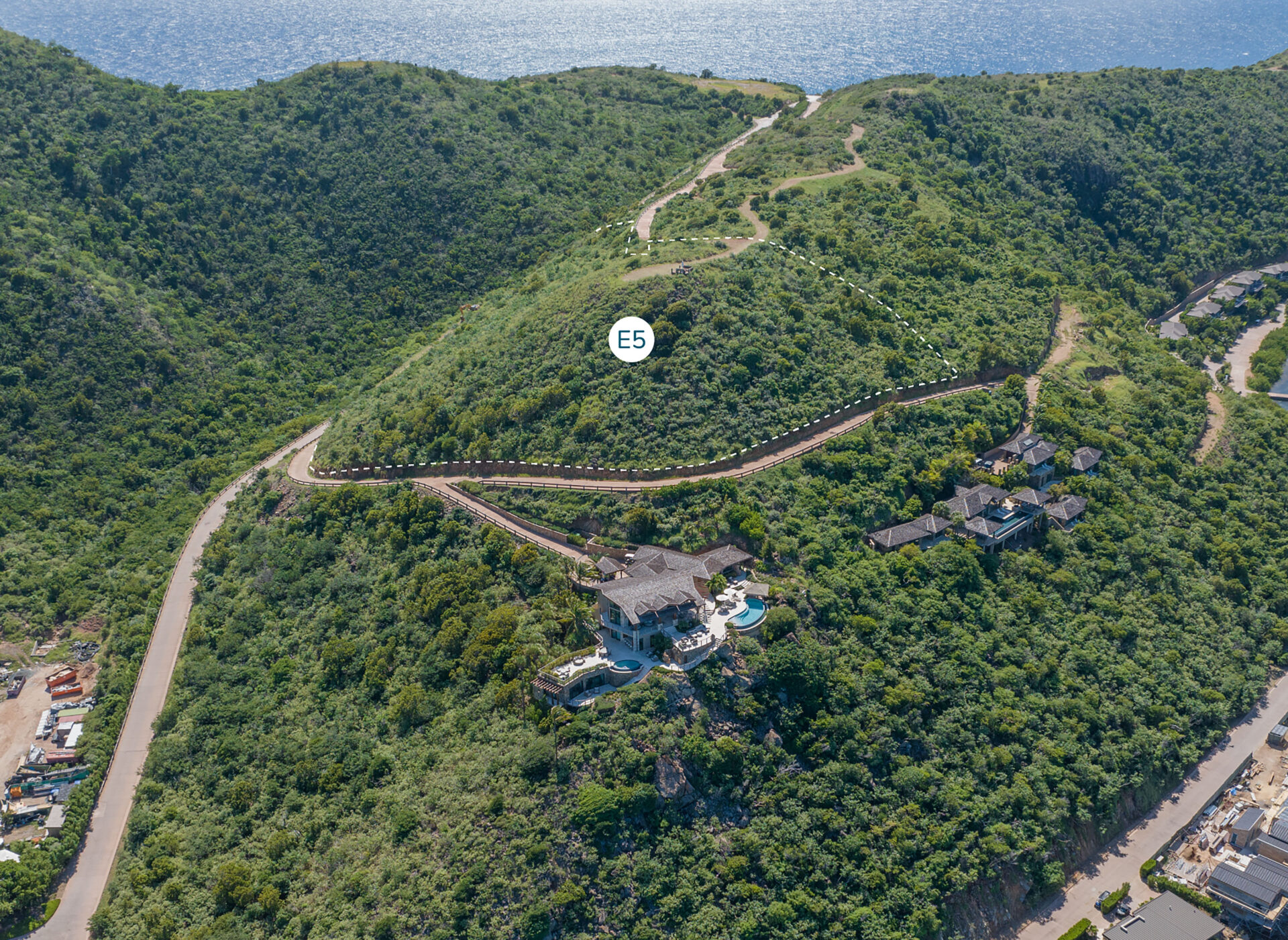 Aerial view of a lush, green hillside with several winding roads leading to an isolated luxury villa with a private pool. The villa prominently sits on the slope, overlooking the ocean in the background. Marked as "E5" in a white circle, this Estate Homesite offers unparalleled privacy and scenic beauty.