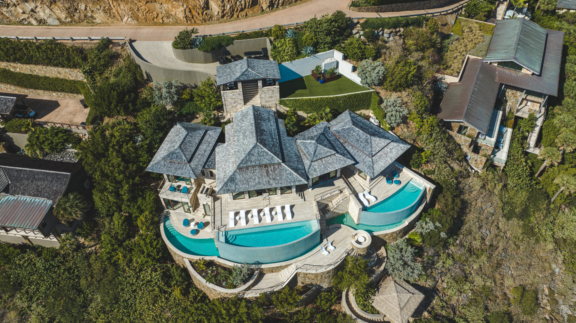 Aerial view of Paradise Villa, a luxurious hillside mansion with multiple four-sided pitched roofs, expansive terraces, and three infinity-edge swimming pools surrounded by greenery. The home features modern design with a backdrop of rugged terrain and winding roadways.