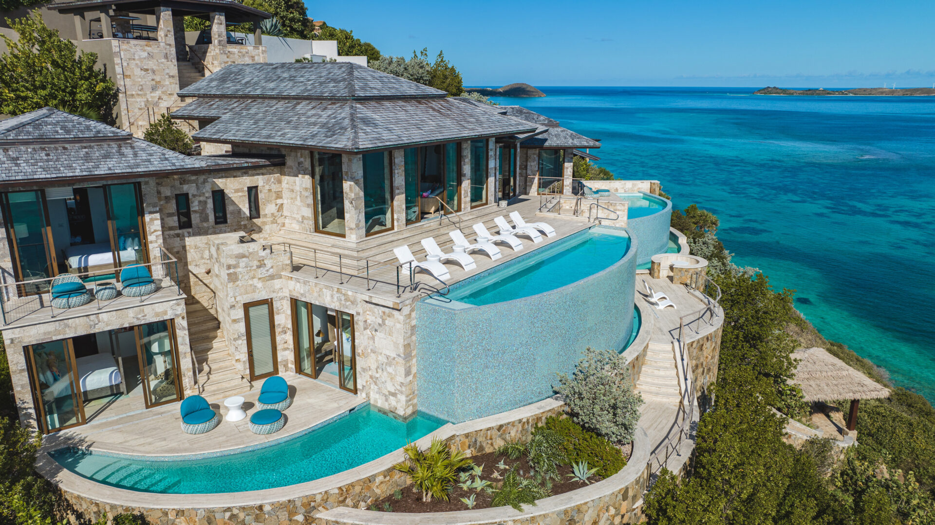 A luxurious beachfront villa, aptly named the Edge of Paradise, boasts stone walls and large glass windows overlooking a clear blue sea. It features multiple terraces, an infinity pool with lounge chairs, and lush greenery. The sky is clear, enhancing the tranquil and scenic ocean view.