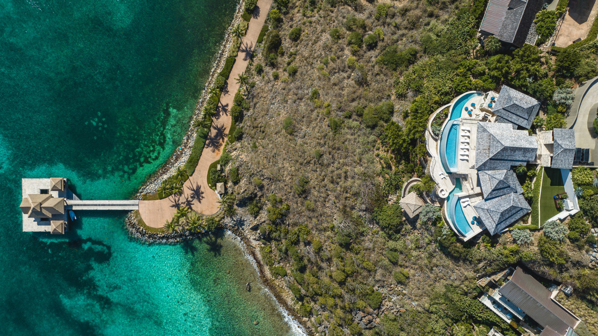 Aerial view of a luxury seaside Villa, aptly named Edge of Paradise, with a pool, surrounded by lush greenery and located next to a clear turquoise ocean. A pathway leads to a wooden pavilion over the water. The property and coastline are beautifully integrated with the natural landscape.