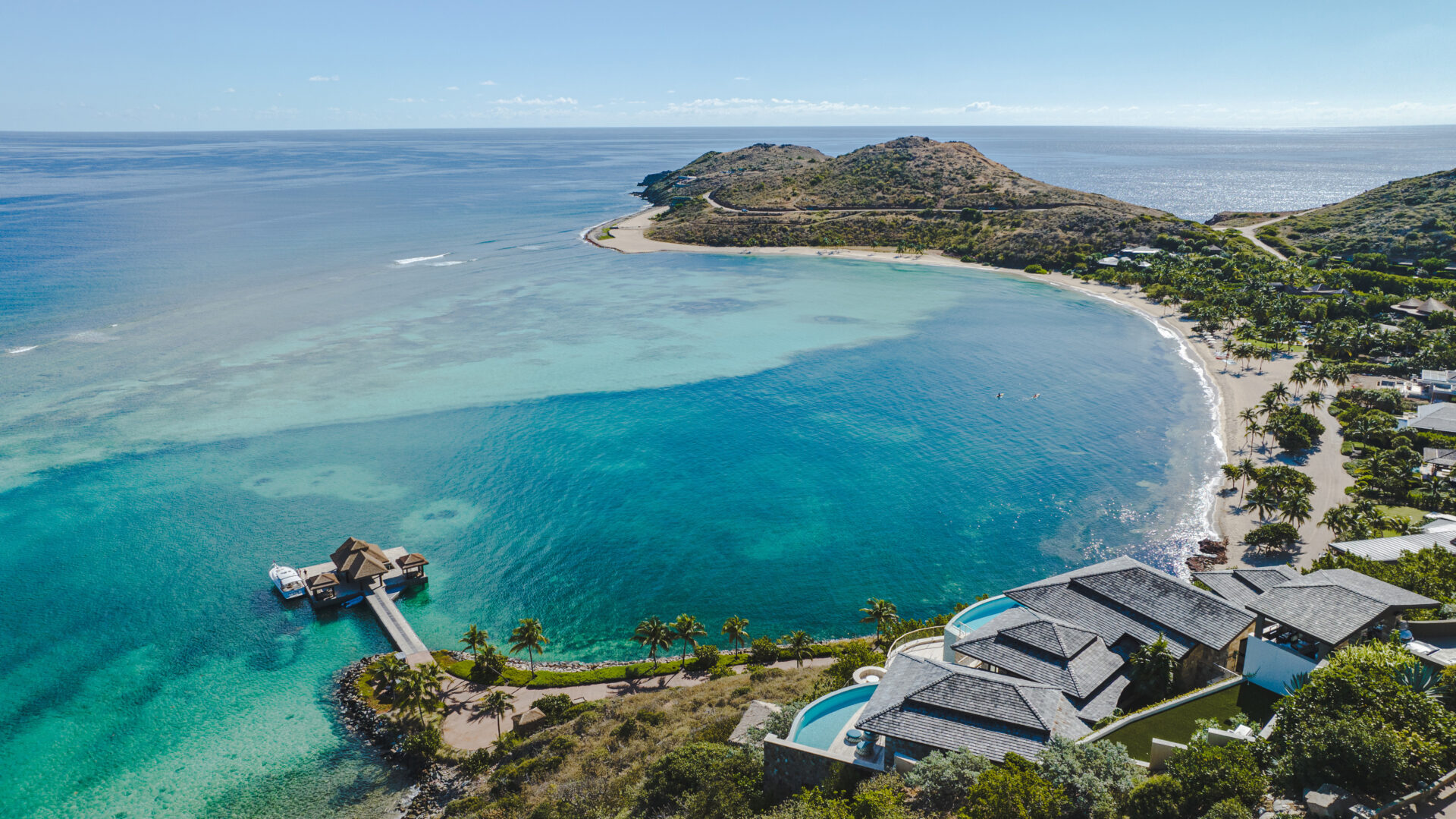 Aerial view of the Edge of Paradise, featuring a luxurious beachfront property with an infinity pool, a wooden pier extending into the turquoise sea, and a sandy beach curved around a hilly peninsula. Clear blue skies and a calm ocean complete the serene scene.