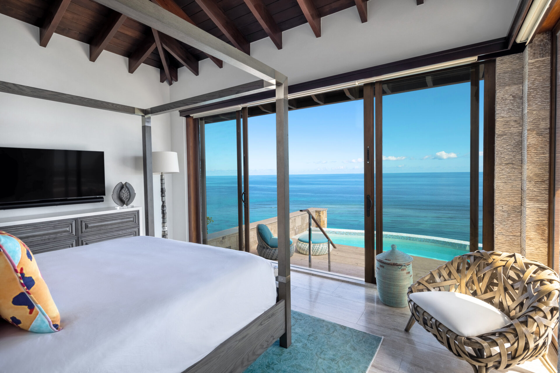 A bright, airy bedroom at Paradise Villa with ocean views features a four-poster bed with white bedding, a large flat-screen TV on a wooden stand, a cozy chair with a woven design, and floor-to-ceiling windows and door leading to a balcony overlooking the clear blue sea.