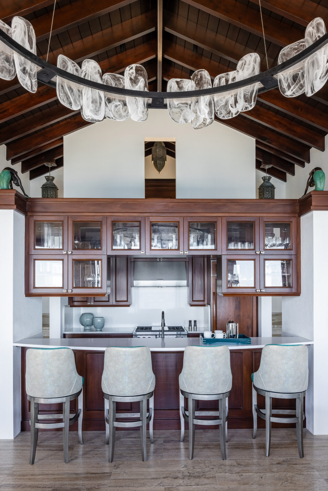 The Edge of Paradise Villa features a stylish home bar with dark wood cabinets and glass fronts, a counter lined with four cushioned bar stools, and a modern chandelier with glass elements. The high wooden beam ceilings enhance the space, while decorative items are elegantly displayed atop the cabinets.