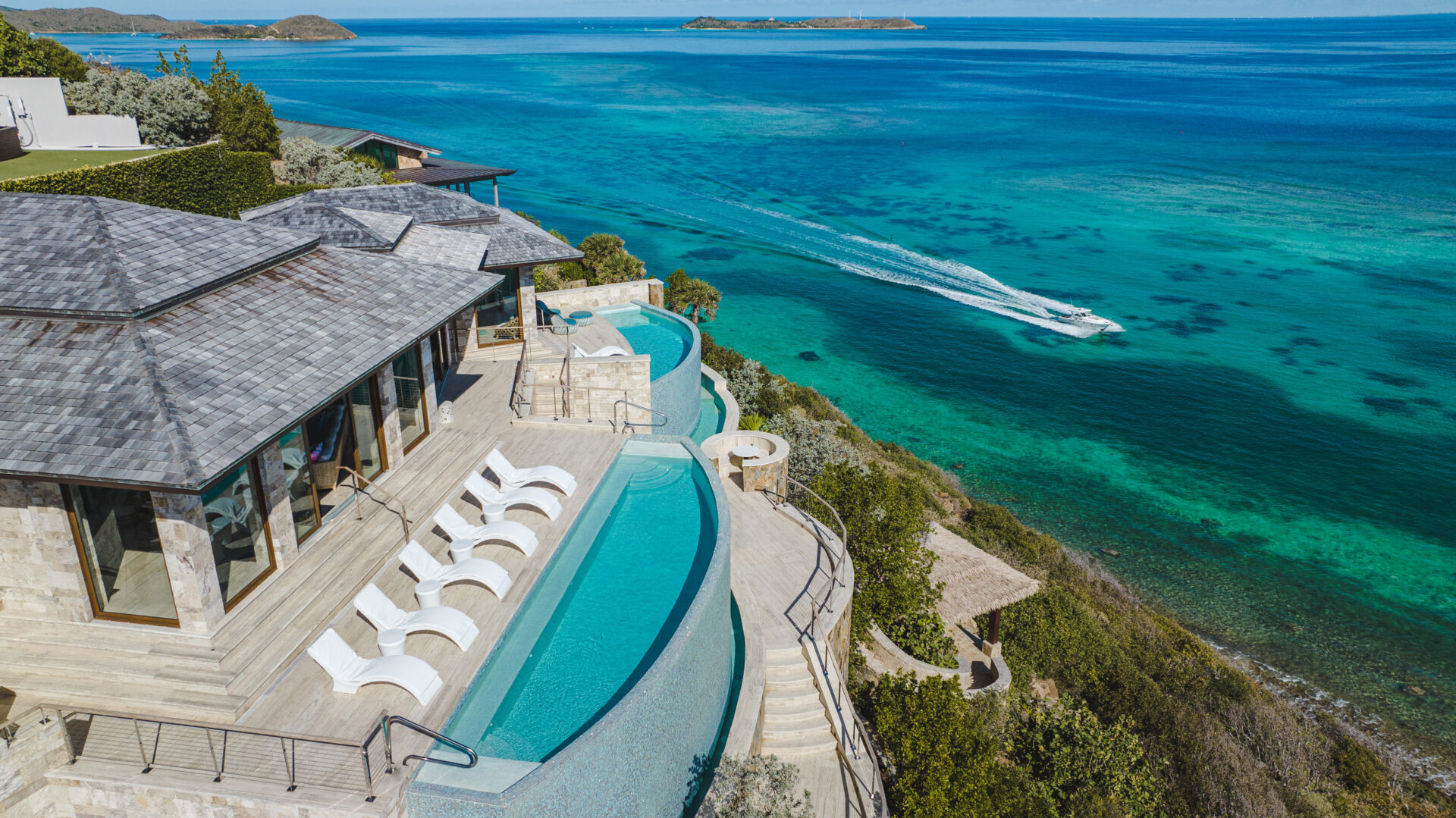 A luxurious villa at the Edge of Paradise, overlooking a turquoise ocean. The villa features an infinity pool with several poolside loungers, blending perfectly with the sea view. A speedboat cruises in the distance, leaving white trails in the clear blue water. Surroundings are lush and green.