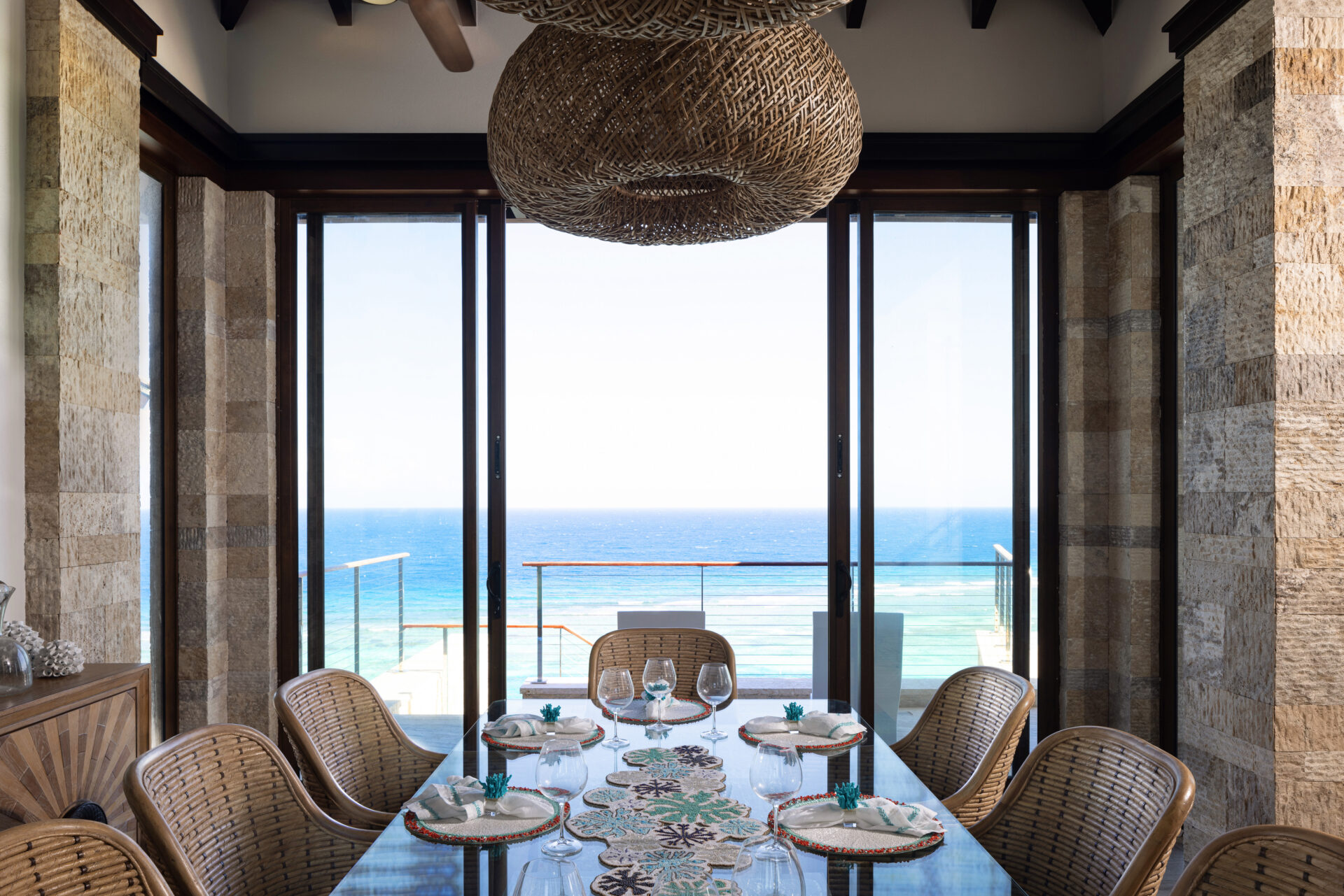 A dining room at Paradise Villa features a large glass table set for a meal, surrounded by wicker chairs. The room has large glass doors that offer a stunning view of the ocean at the edge of paradise. A woven wicker light fixture hangs from the ceiling, adding a natural touch to the modern decor.