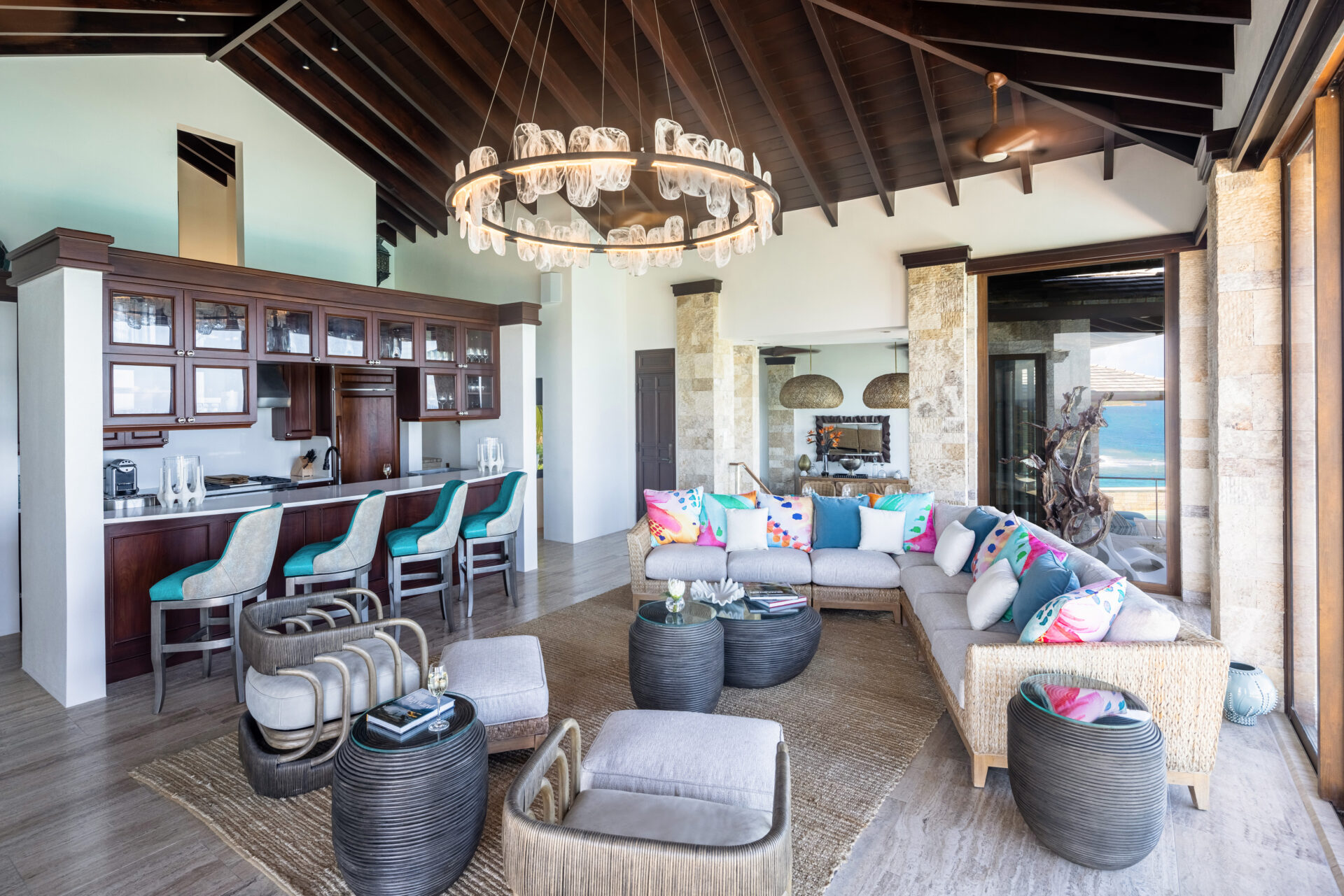 A spacious, modern living room with large windows offering an ocean view at the edge of paradise. The villa features cushioned seating with colorful pillows, a circular chandelier, a bar area with teal stools, contemporary decor, and a high ceiling with exposed dark wooden beams.