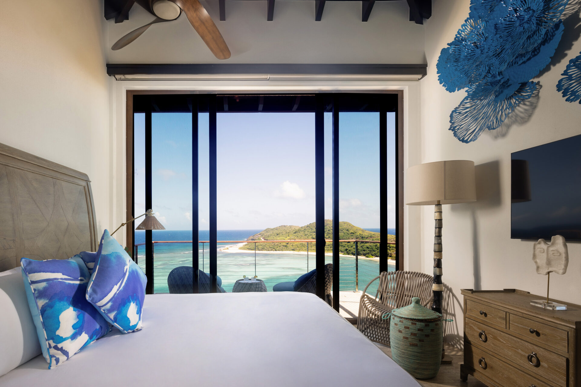 A modern bedroom in the Paradise Villa with a large glass door opening to a balcony offering a stunning ocean and island view. The room features a bed with blue and white pillows, a ceiling fan, a wicker chair, a wooden dresser with a lamp, and blue decorative wall art.