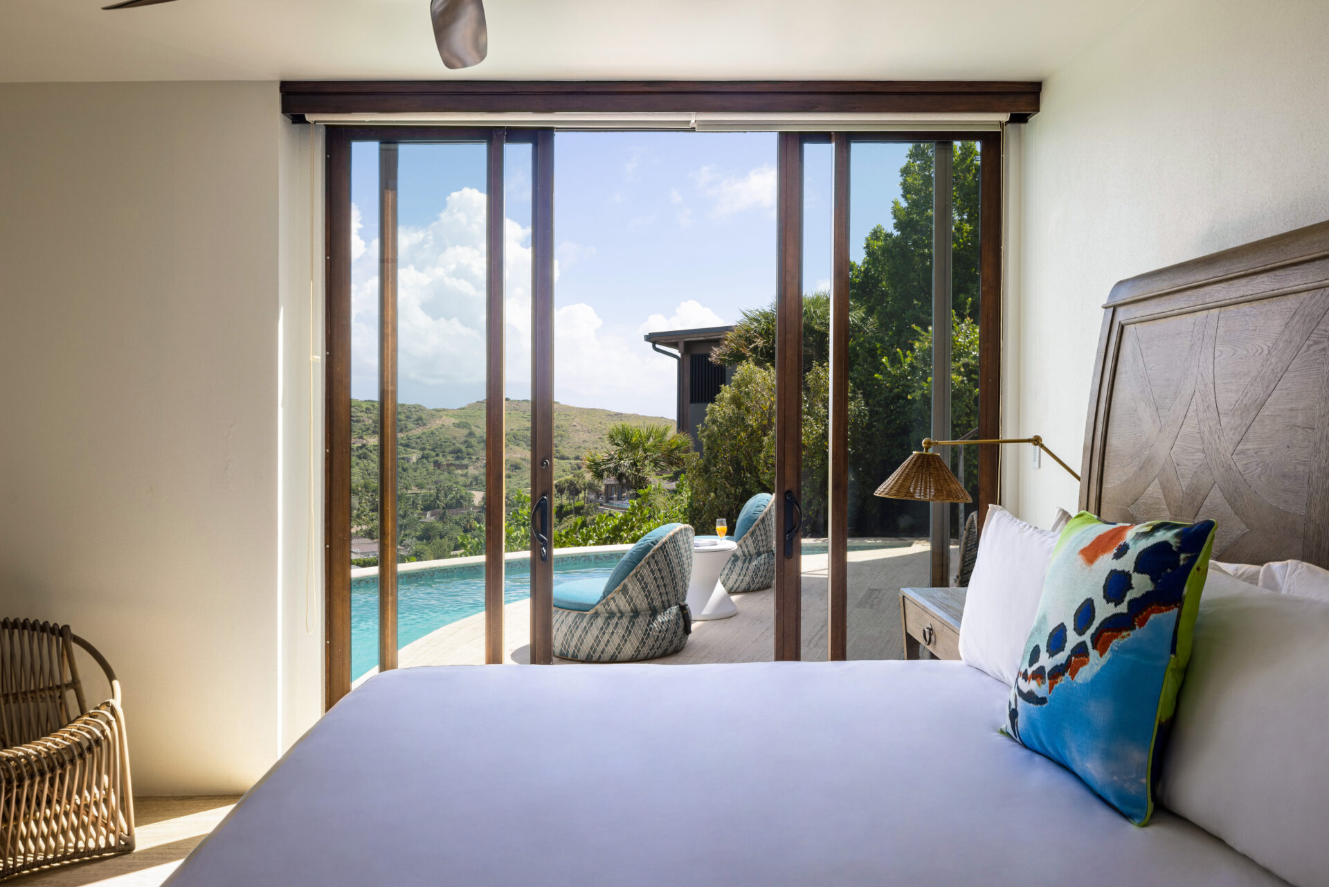 A serene bedroom at the Edge of Paradise villa features a large bed, colorful cushions, and a wicker chair. Sliding glass doors open to a patio with lounge chairs, a pool, and scenic hills in the background. The room is bright with natural light, offering a modern and relaxing ambiance.