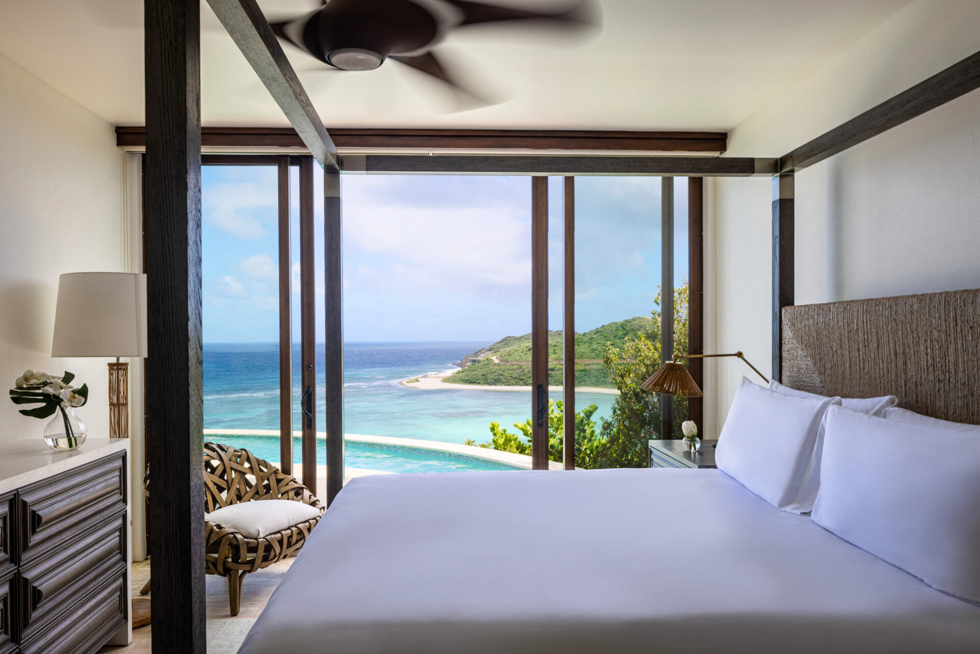 A serene bedroom in Paradise Villa features a large canopy bed, white linens, and modern decor, opening to a stunning ocean view through floor-to-ceiling windows. Outside, a turquoise sea meets a lush green coastline. A wicker chair and lamp by the window enhance the tranquil setting at this Edge of Paradise.