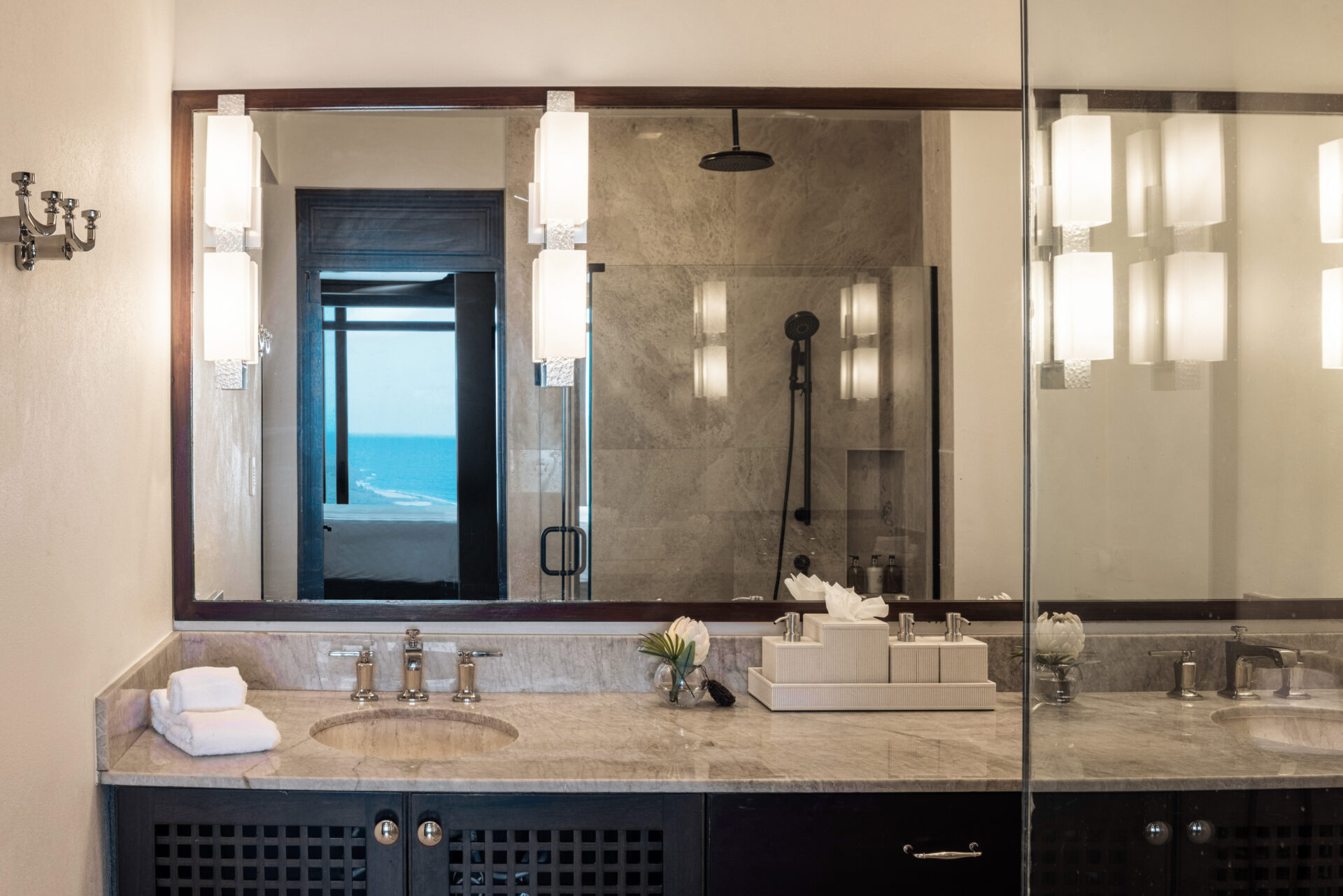 A modern bathroom in Edge Villa featuring a large mirror with mounted light fixtures on both sides. Below the mirror, a marble countertop holds a sink, towels, and a tissue box. In the background, there's a glass-enclosed shower and a window revealing stunning ocean views.