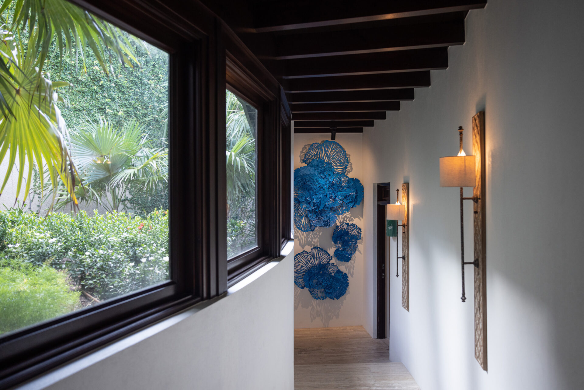 A narrow hallway with a large window on the left side overlooking lush greenery, giving a glimpse of the Edge of Paradise. On the right wall, two wall lamps illuminate a vibrant blue abstract art installation displayed at the end of the corridor. The ceiling features exposed beams.