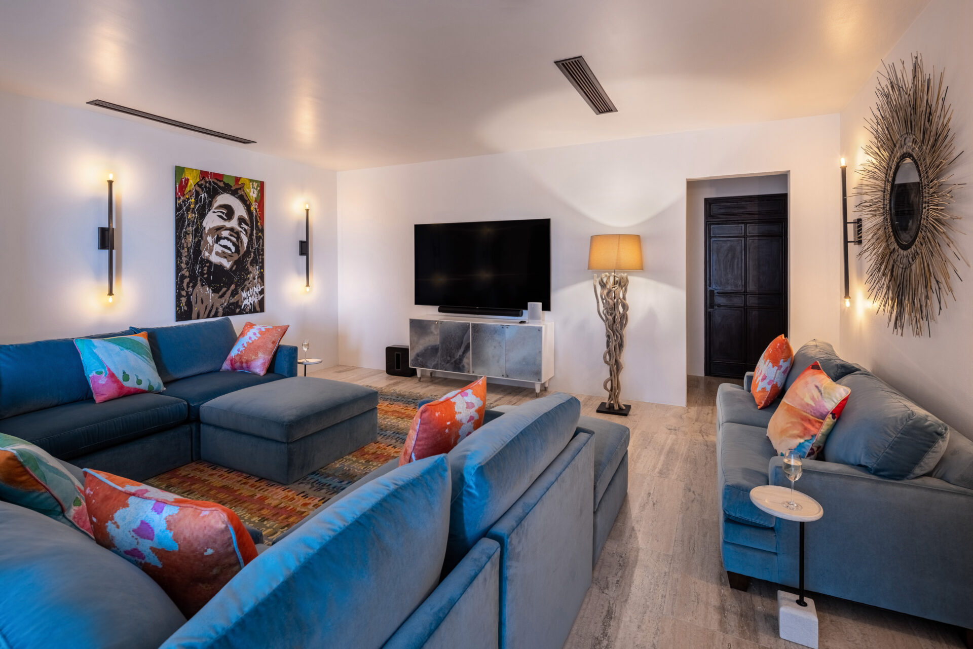 A modern living room at Edge of Paradise features blue sectional sofas and colorful cushions. A large TV is mounted on the wall, a painting of a smiling person graces the left wall, and a tall lamp with a unique twisted base stands near a dark door. The room is warmly lit.