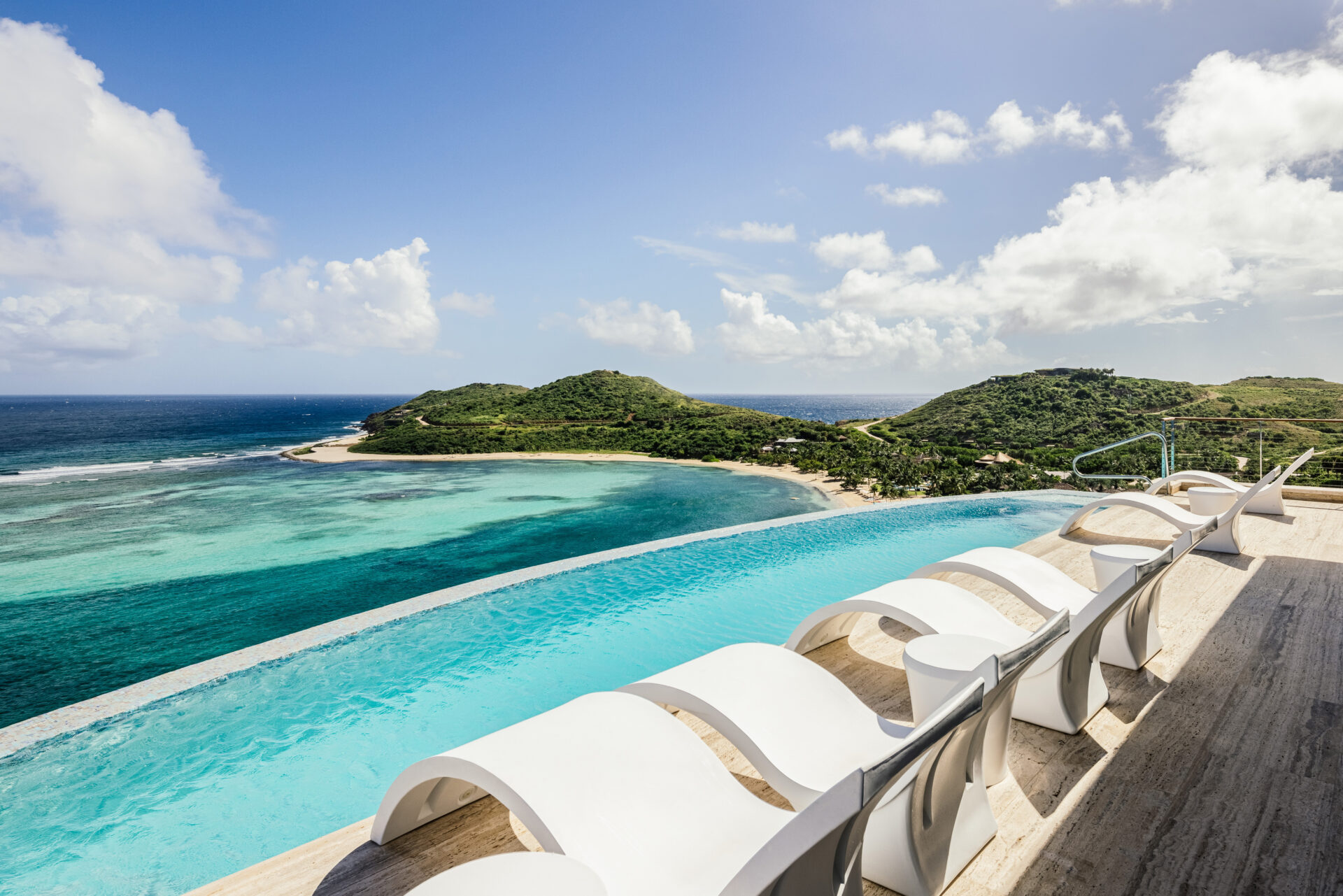 A luxurious infinity pool with sleek white lounge chairs overlooks a stunning tropical coastline at the Edge of Paradise Villa. The crystal-clear water of the pool seamlessly merges with the turquoise ocean below. Green hills and a partially cloudy blue sky complete the serene scene.