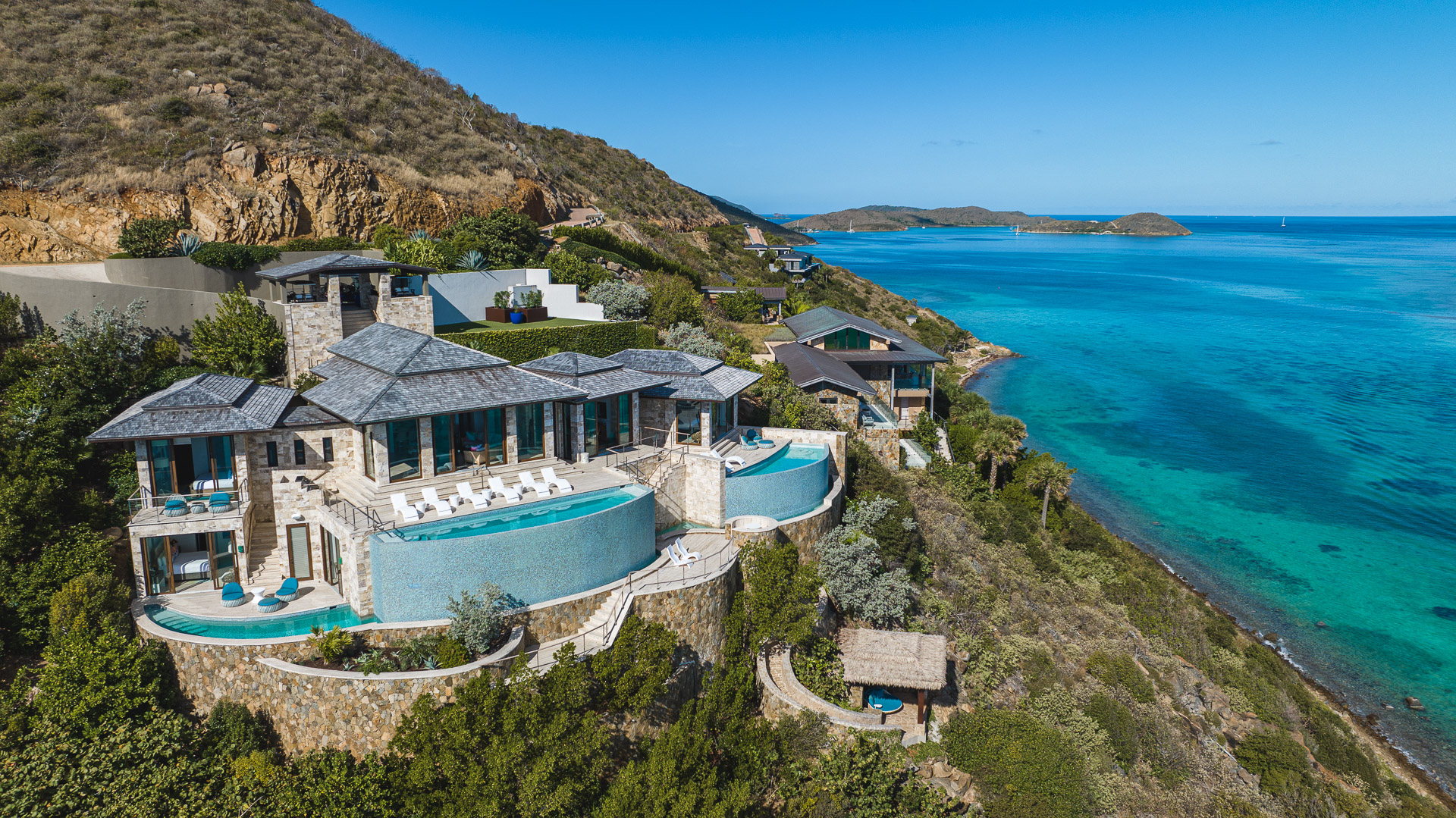 Edge of Paradise Villa, a luxury retreat perched on a hillside, overlooks a turquoise-blue ocean. The contemporary stone and glass residence features a prominent infinity pool, lush greenery surrounds the property, along with neighboring villas on the slope and clear blue sky overhead.