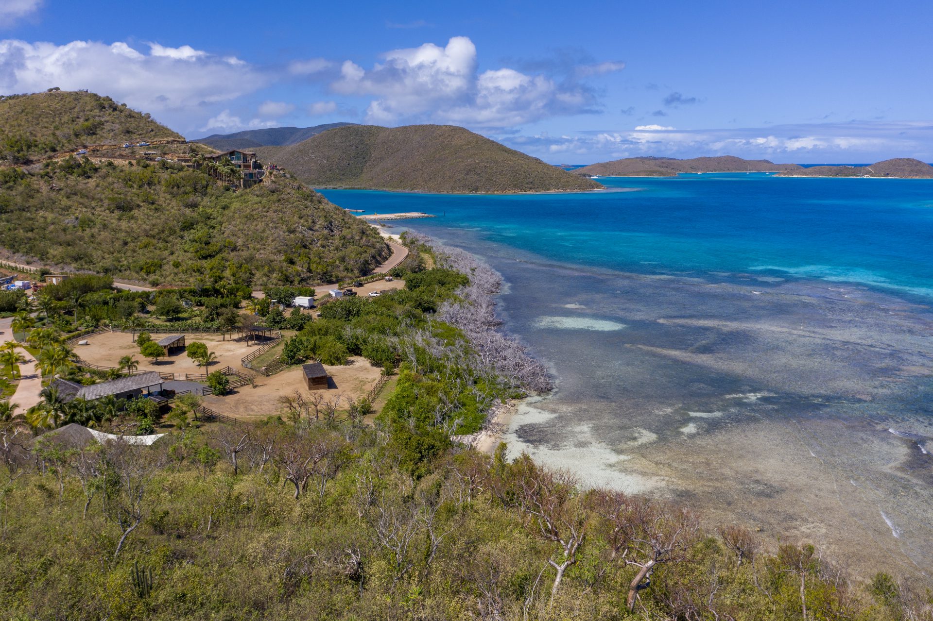 Aerial view of a tropical coastline featuring clear turquoise waters and a coral reef. The landscape includes lush green hills, scattered houses, and winding roads, with E19 marking an ideal estate homesite. The sky is partly cloudy with sunlight illuminating the vibrant scenery.