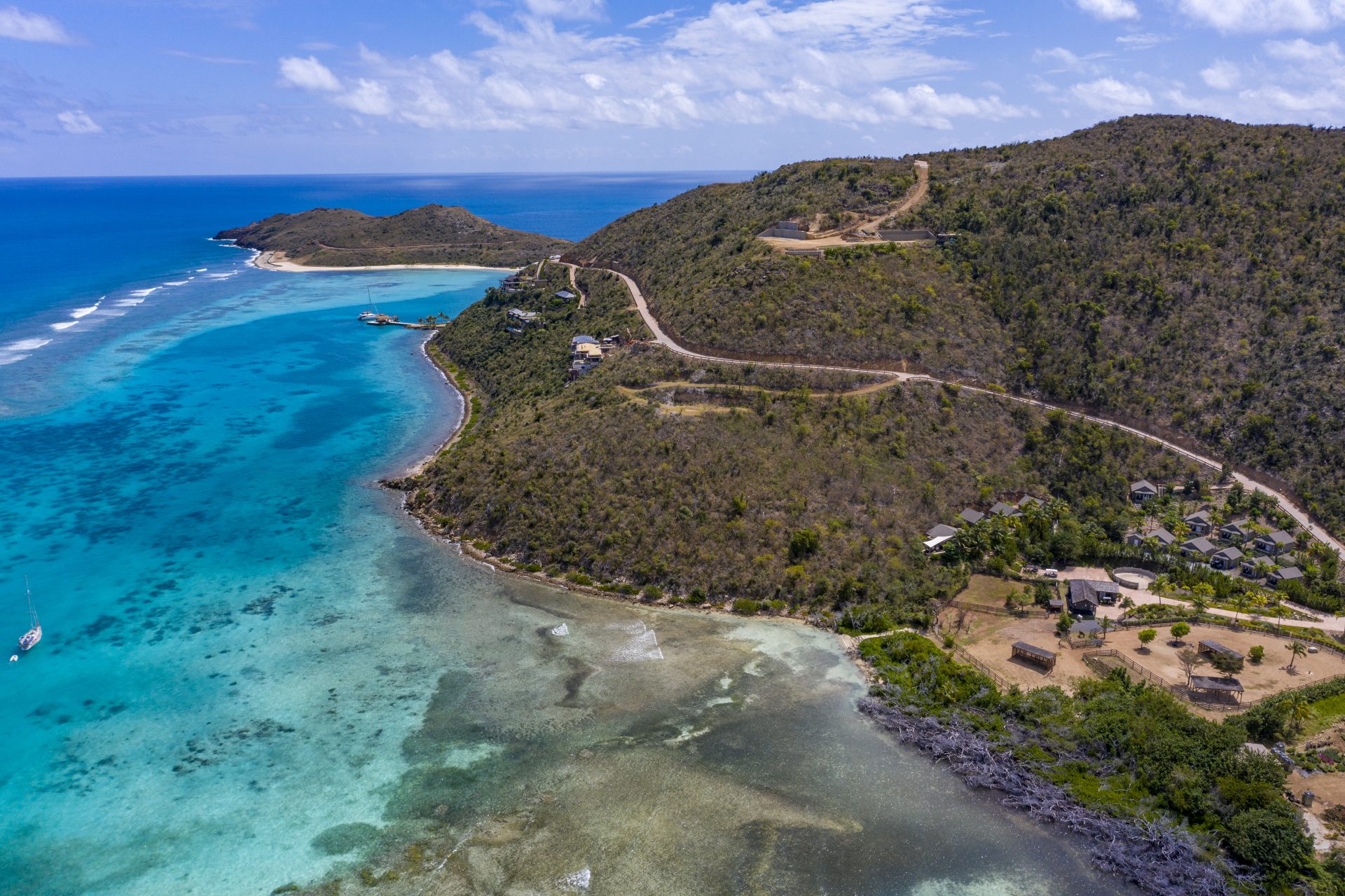 Aerial view of a scenic coastal landscape with turquoise waters, hilly terrain, and a winding road. The coastline features estate homesites, small buildings, lush greenery, and sandy beaches. An anchored boat is visible in the clear, shallow waters near the shore.