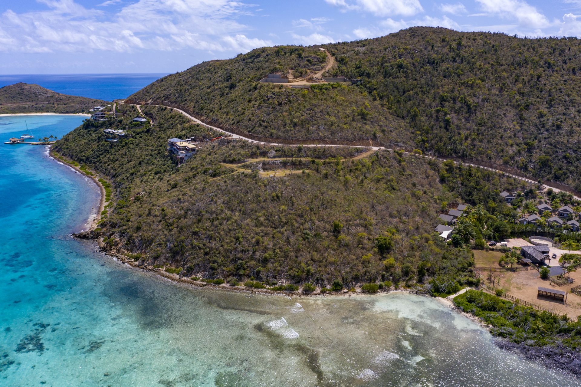 Aerial view of a coastal landscape with turquoise blue waters, rugged hills, and sparse vegetation. A winding road traverses the hillside, passing several buildings and leading to Estate Homesite E19 near the shore. Clear sky with a few scattered clouds.