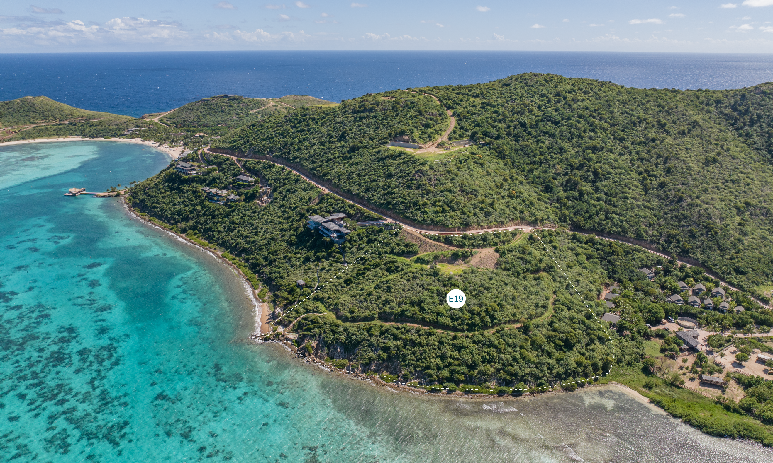 Aerial view of a lush, green island with a winding road leading to a hilltop. Crystal-clear turquoise water surrounds the island, showcasing a small bay on the left. A marker labeled "E19" designates an exclusive homesite within the hillside vegetation.