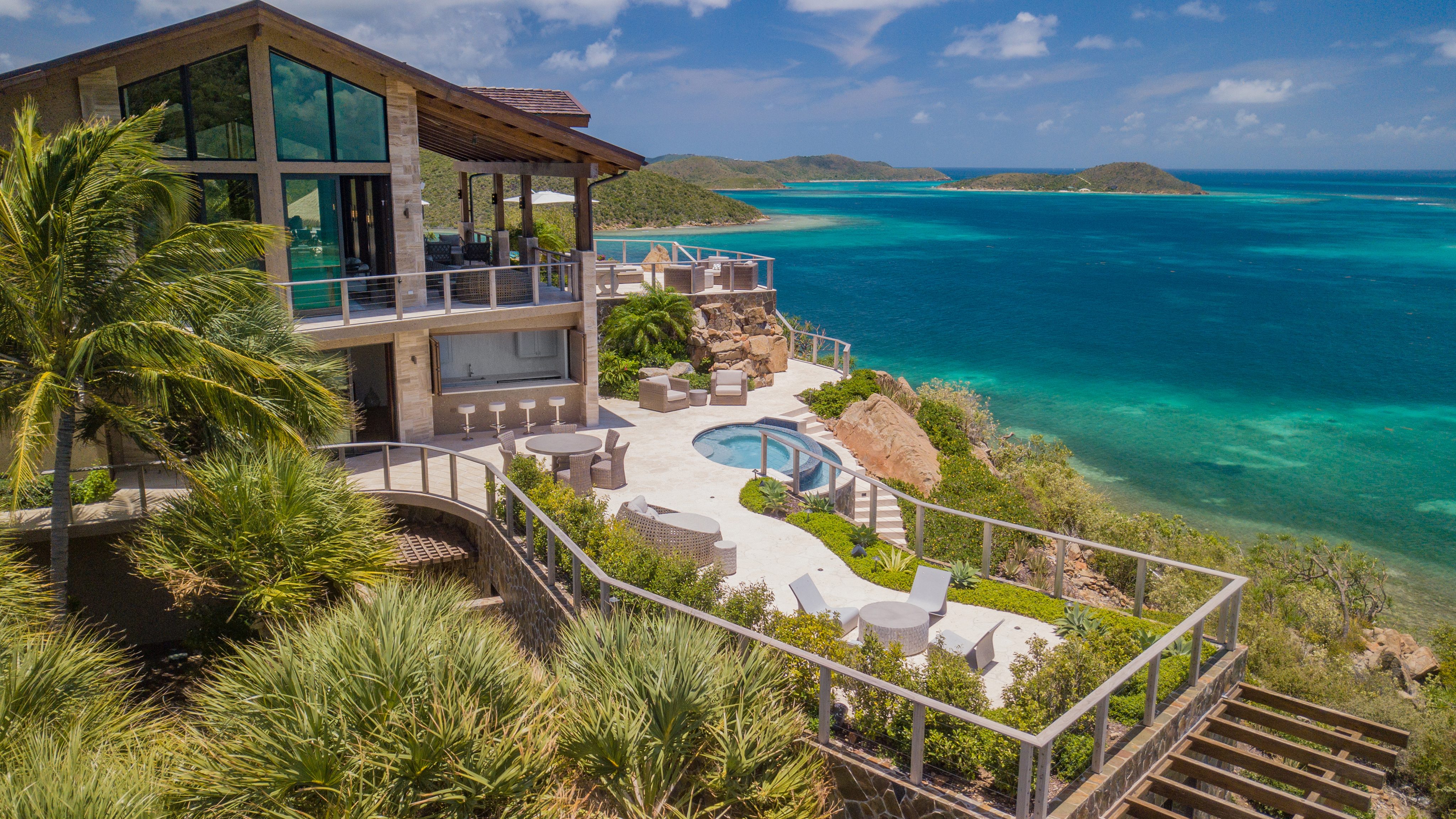 A modern seaside villa with large glass windows sits atop a cliff, overlooking a turquoise ocean and lush green islands. The villa features a spacious patio area with a round swimming pool, lounge chairs, and outdoor seating surrounded by tropical plants, resembling an art gallery of nature’s finest views.