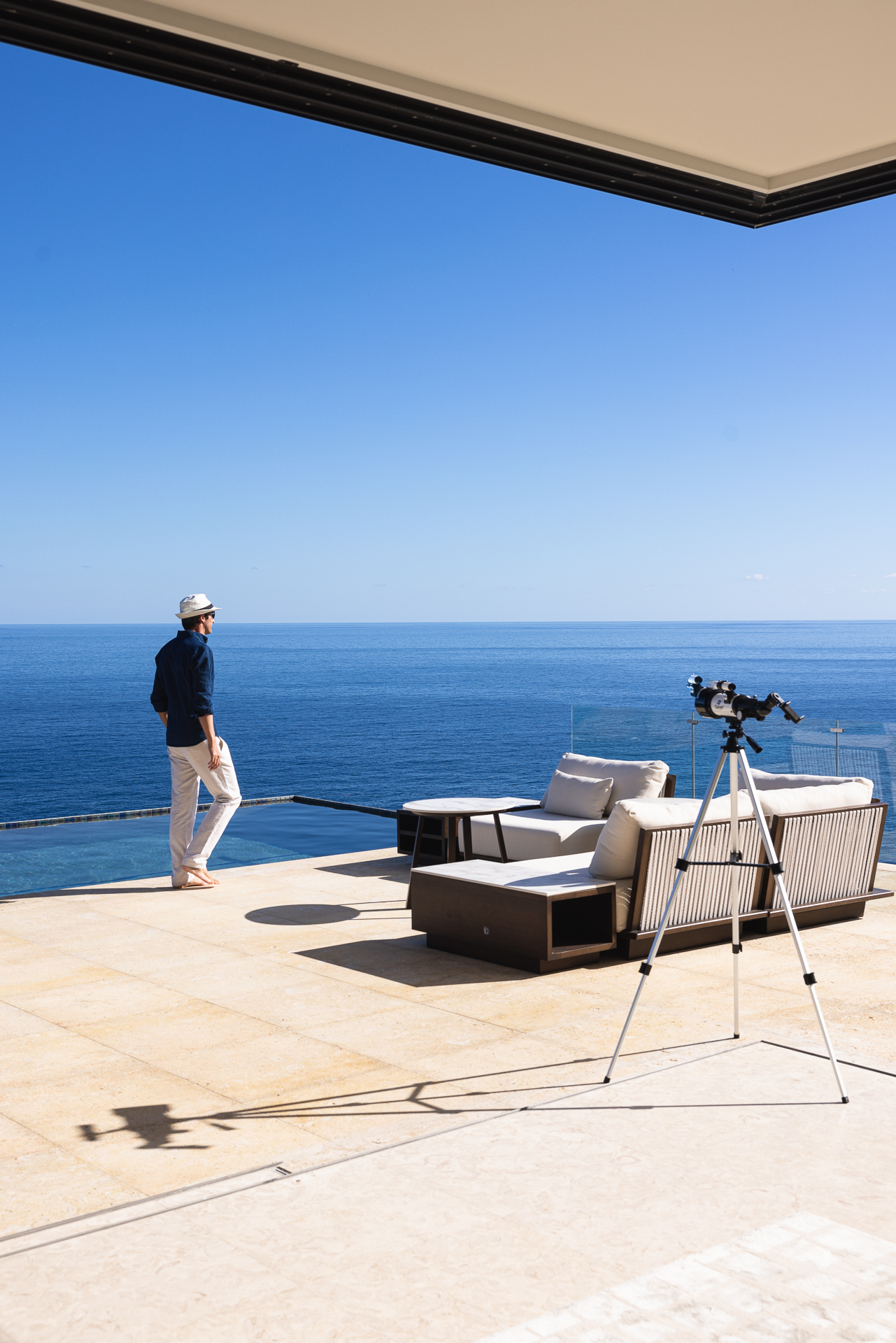 A person stands on a spacious terrace overlooking the ocean under a clear blue sky. On the terrace are two cushioned lounge chairs and a telescope mounted on a tripod, oriented towards the horizon. The scene exudes tranquility and luxury, embodying an upscale lifestyle.