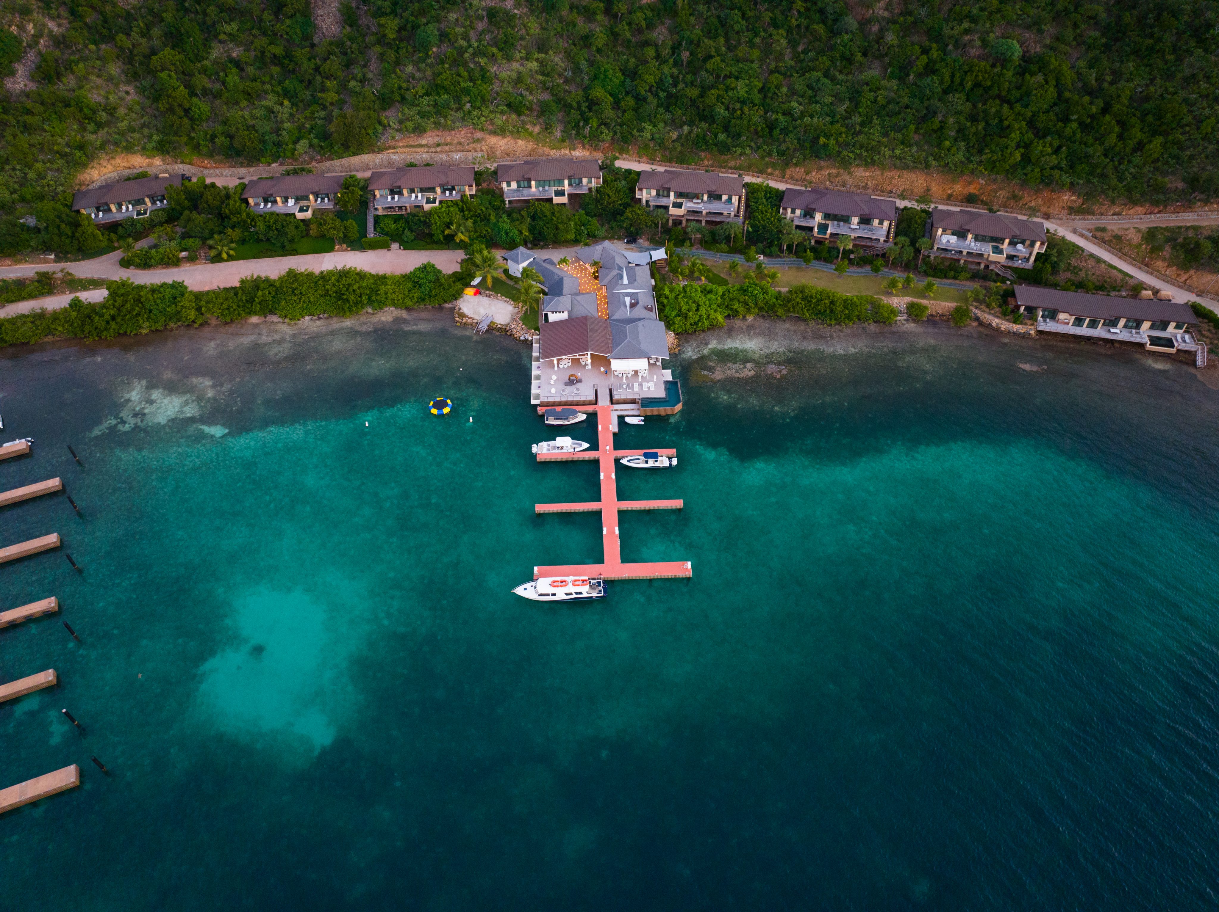 Aerial view of a waterfront coastal resort featuring several buildings along the shoreline. A central pier extends into the turquoise water, with boats docked along it in the marina. Surrounding greenery and a mountainous backdrop complete the picturesque scene.