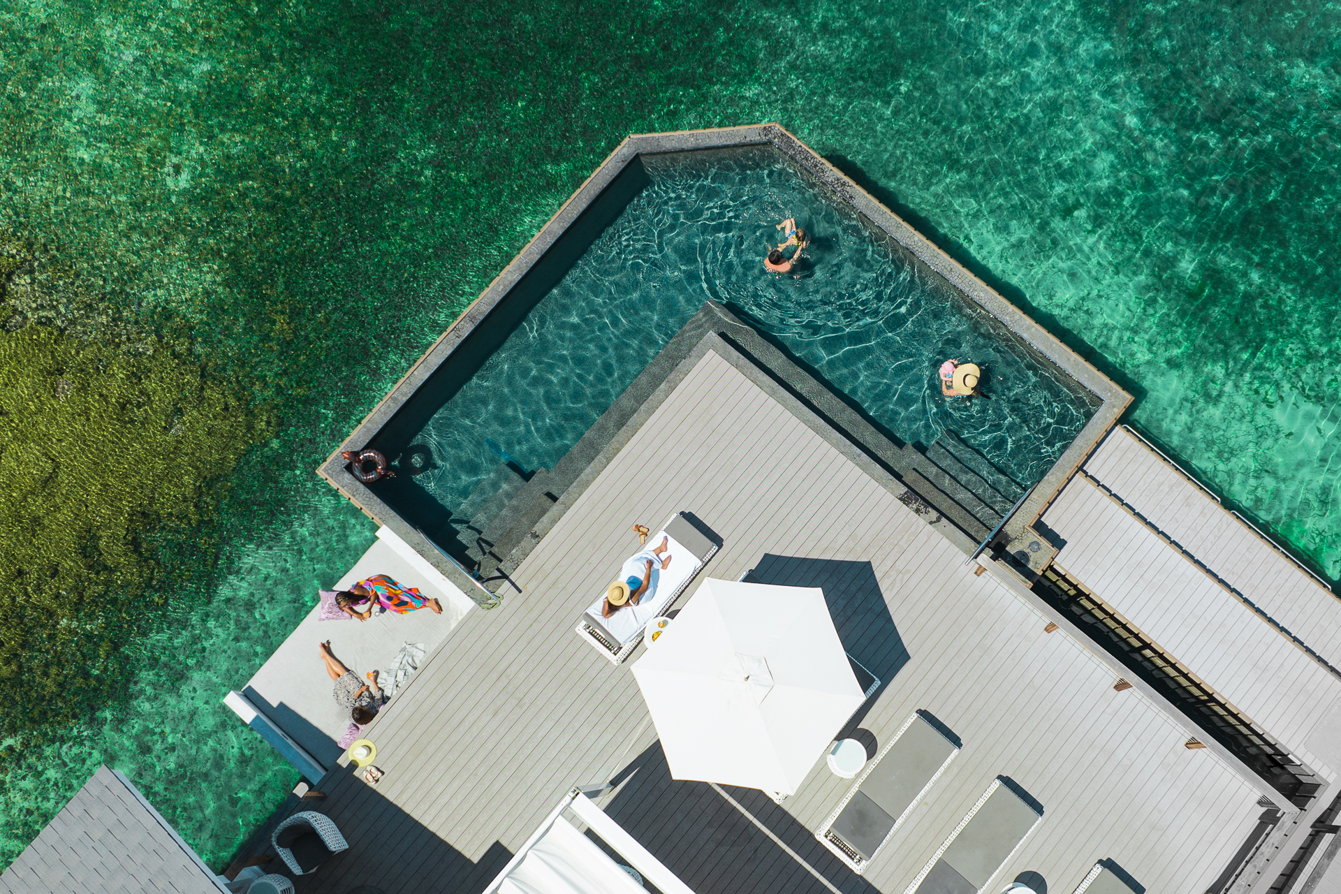 Aerial view of a deck at Marina Village with people sunbathing and swimming. The deck features lounge chairs, umbrellas, and a pool adjoining the turquoise ocean. Clear water reveals underwater details, and a vibrant towel is spread out on one side of the deck.