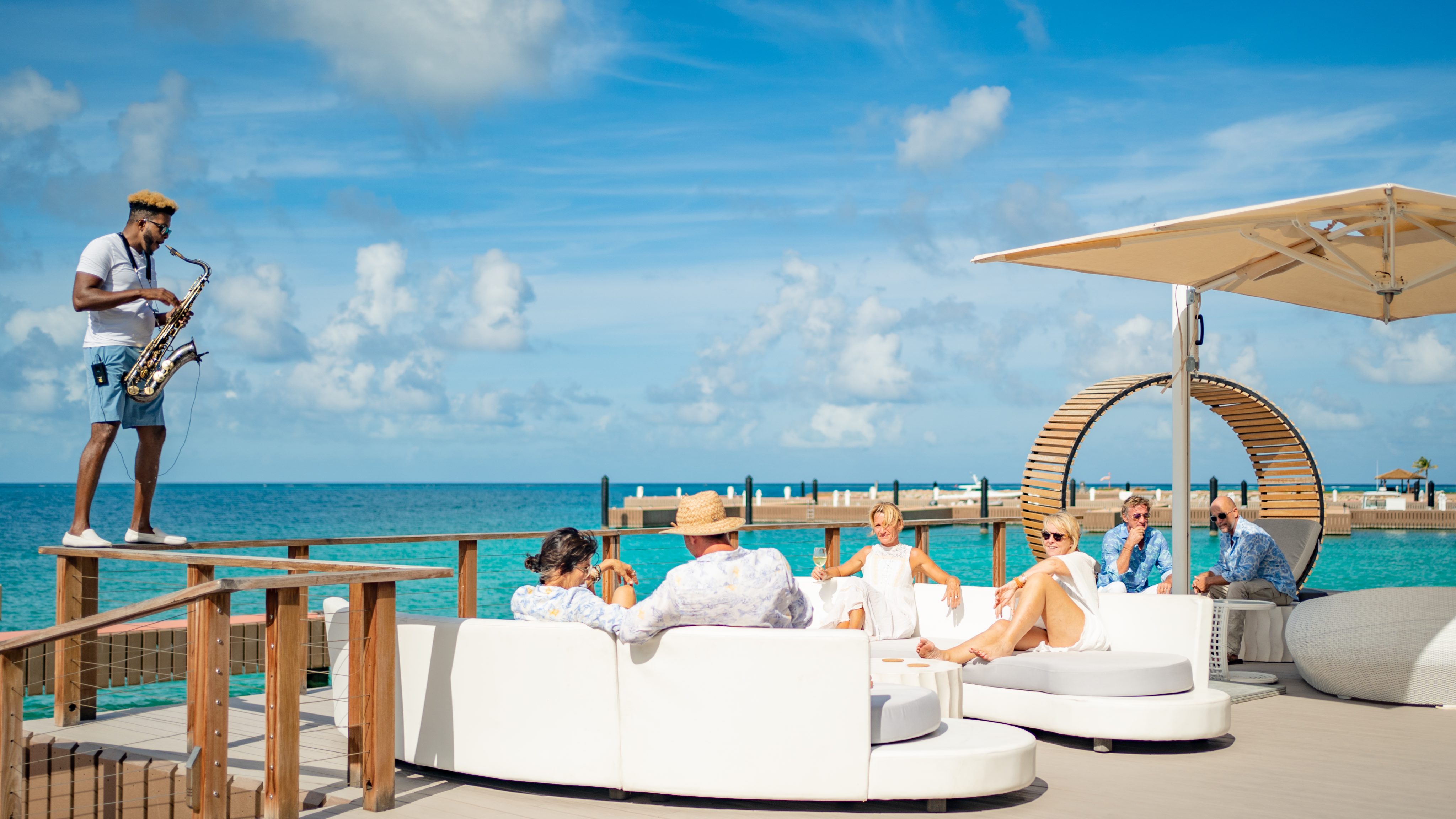 A saxophonist in casual attire performs on a wooden platform beside turquoise ocean waters at the marina. Several people relax in white lounge furniture under a large umbrella, enjoying the sunny day and ocean view. The sky is partly cloudy, enhancing the serene ambiance.